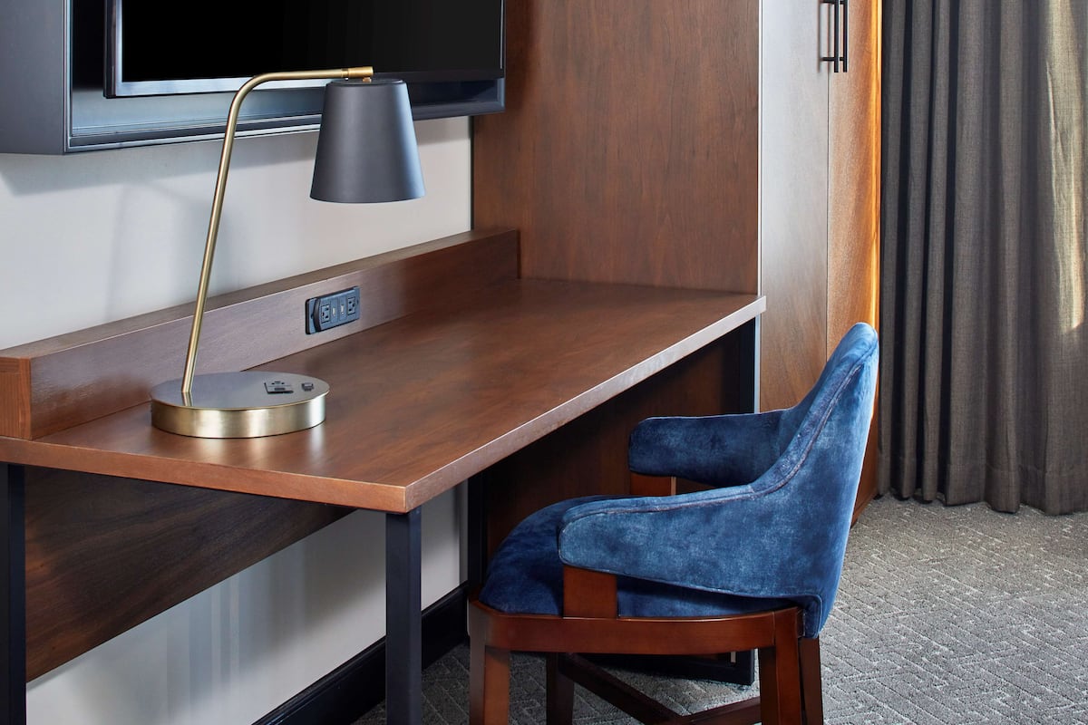 A hotel room with a wooden desk, a blue chair, a TV mounted on the wall, and a power outlet.