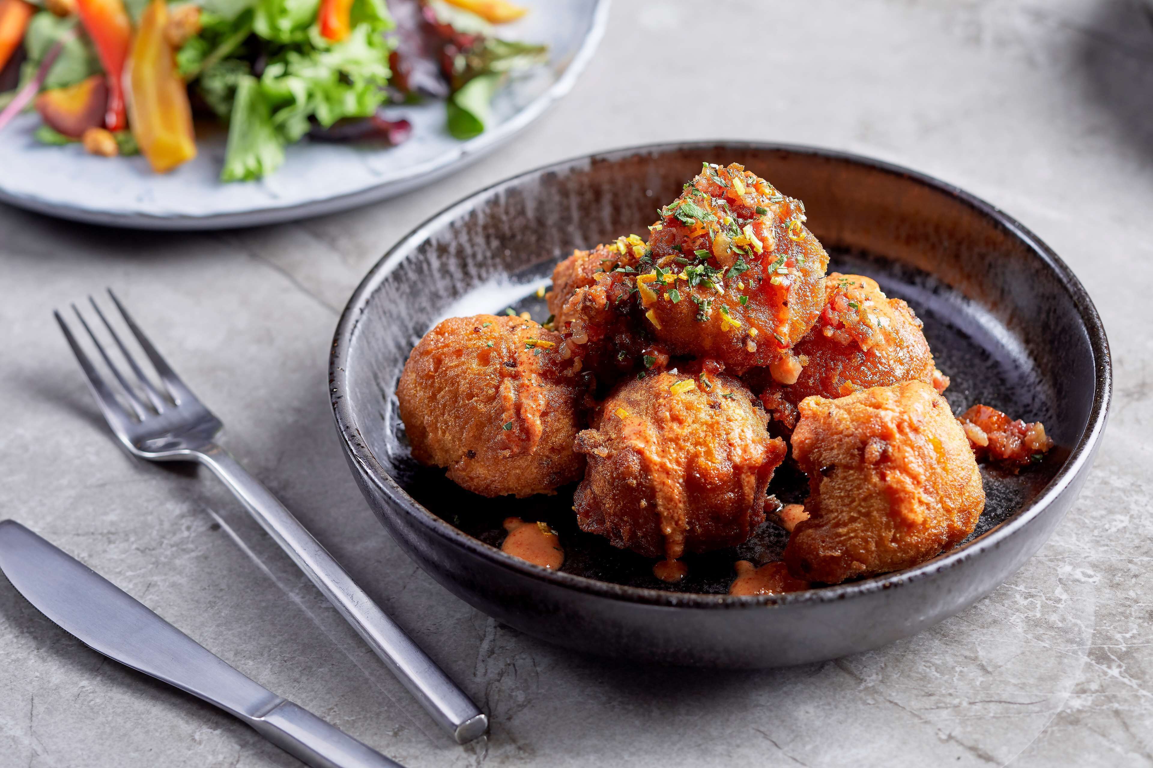 A bowl of fried chicken balls garnished with herbs, accompanied by a fork and knife on a gray surface.