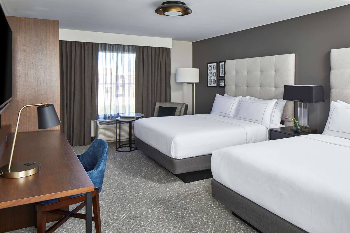 A hotel room with two beds, a desk, a chair, a TV, and a window with curtains.