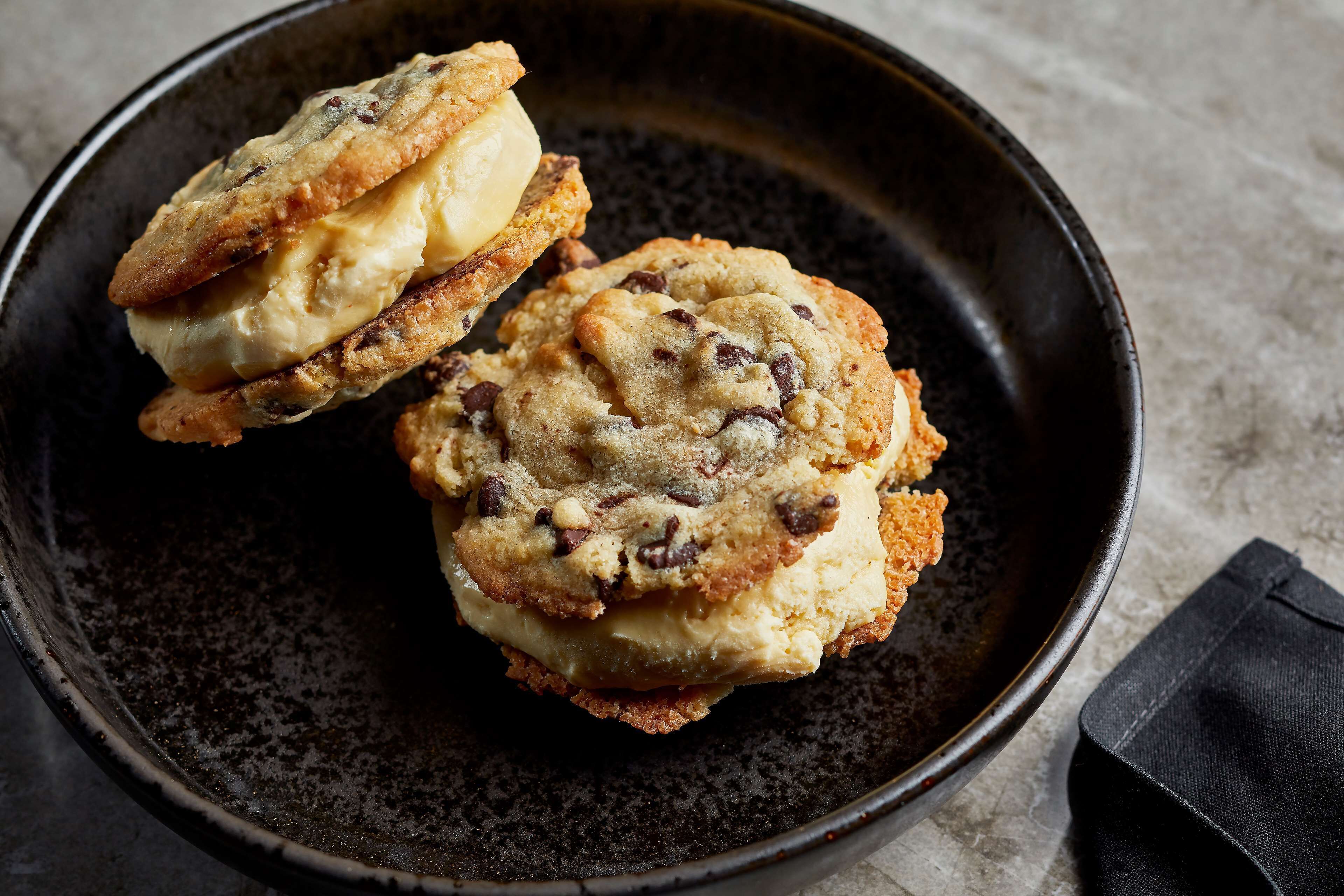 Two chocolate chip cookies with ice cream on a dark plate.