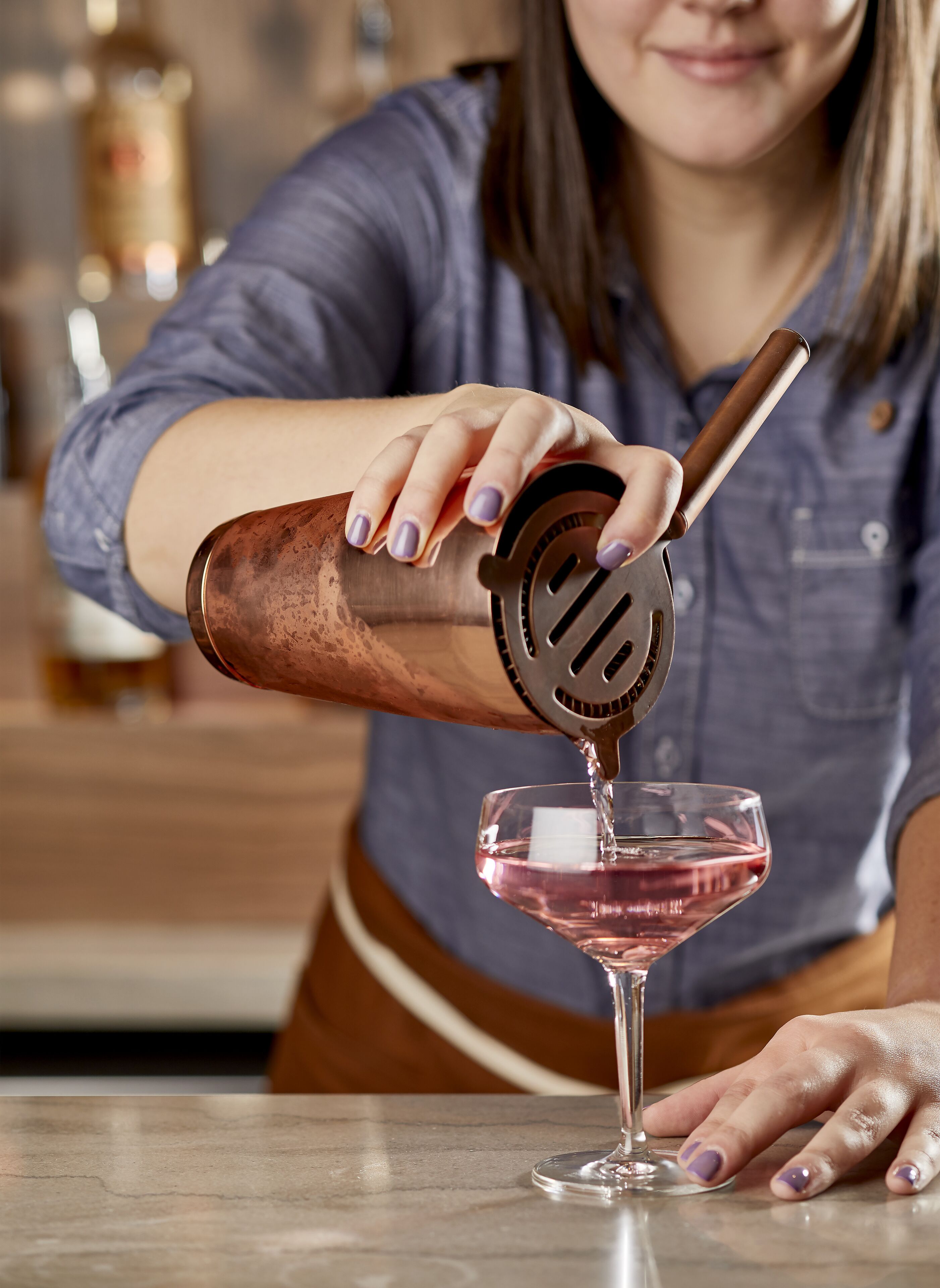 Una persona vertiendo un líquido rosado desde un coctelero de cobre en una copa de martini.