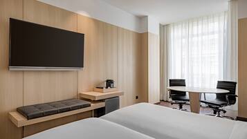 Habitación de hotel con una cama grande, un escritorio con silla, un pequeño rincón de descanso, televisión y ventana con cortinas.