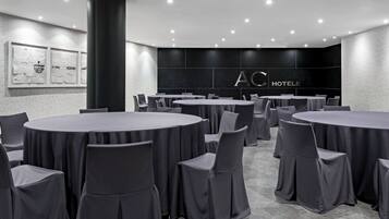 Una sala conferenze con diversi tavoli rotondi e sedie, un'imponente colonna e una parete con il logo AC HOTELS.