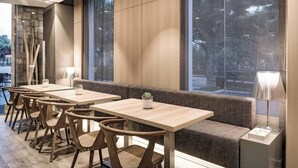 A modern dining area with wooden tables and chairs, a cushioned bench, and large windows offering a view of trees.