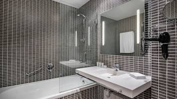 Un baño moderno con bañera de hidromasaje con mampara de vidrio, un tocador blanco con espejo y un secador de pelo negro montado en la pared.