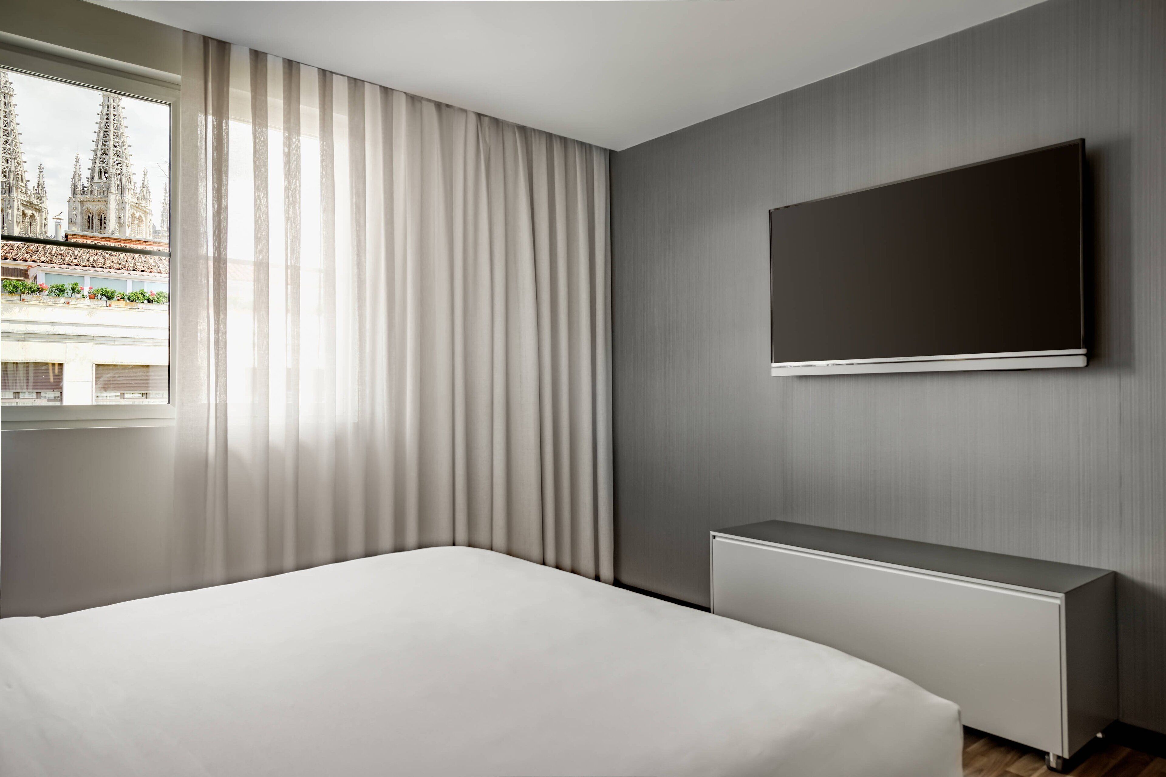 A modern hotel room with a large bed, a flat-screen TV mounted on the wall, and a window with sheer curtains.