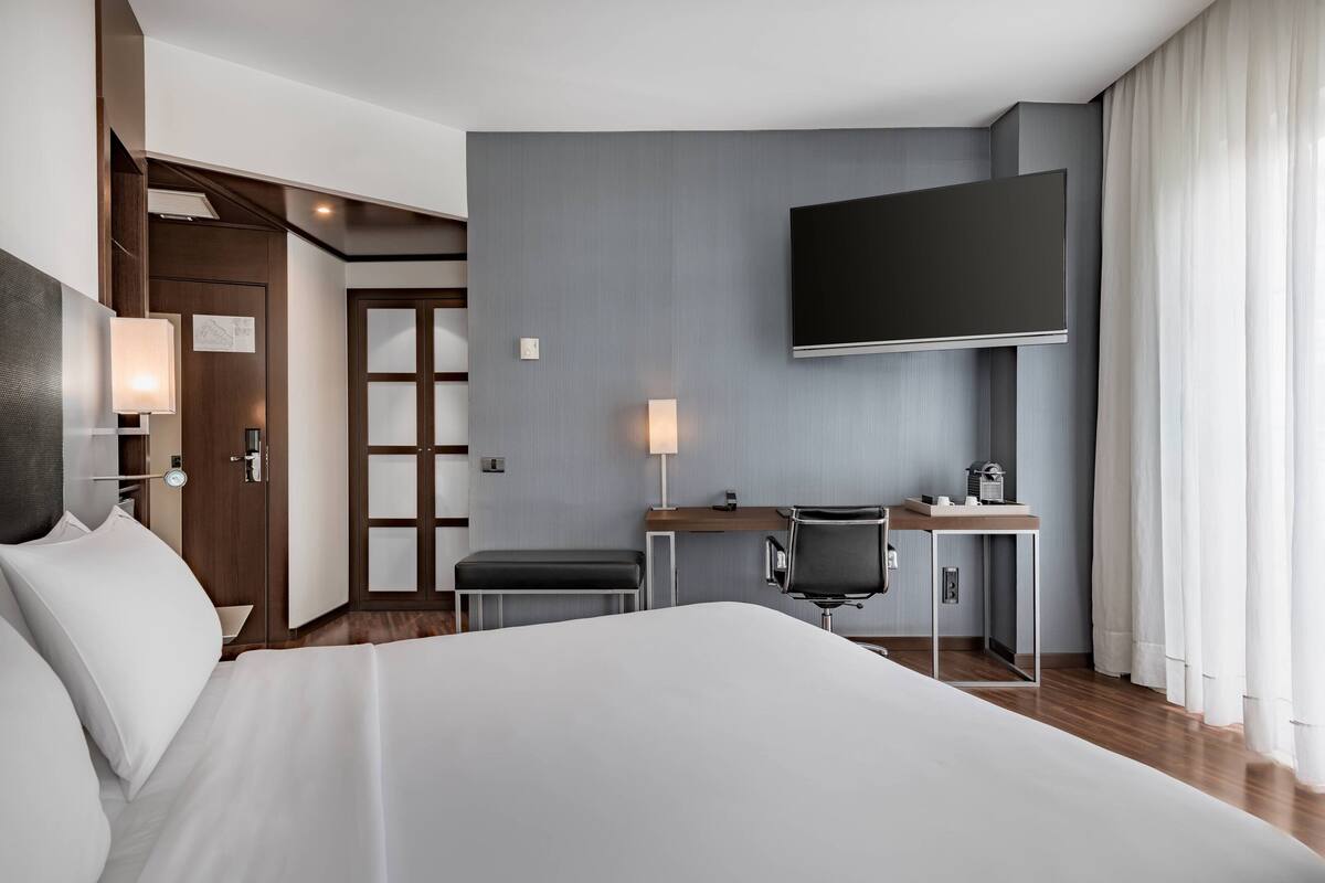 Een moderne hotelkamer met een groot bed, een bureau met stoel, een flatscreen televisie en een groot raam met gordijnen.