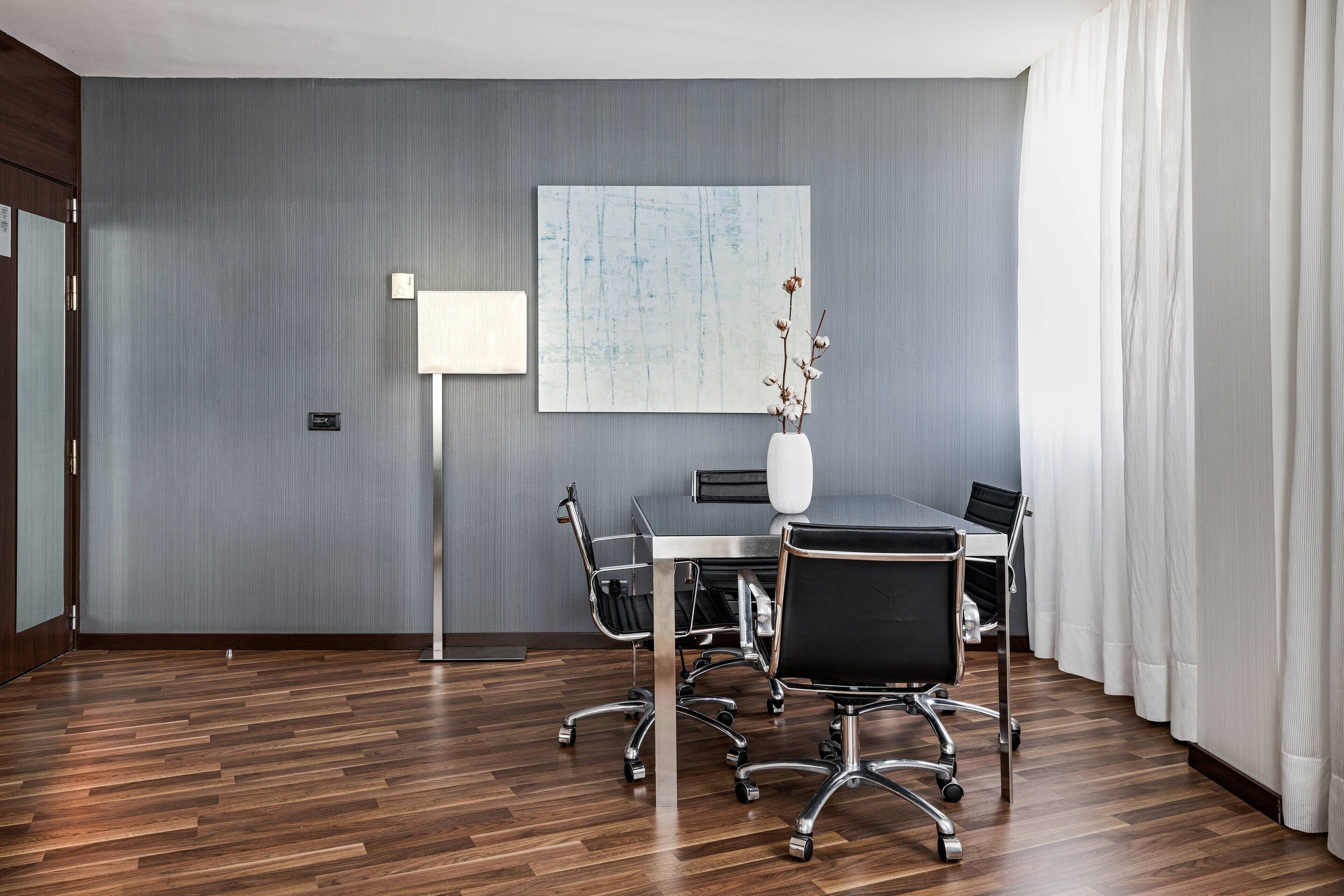 A conference room with a wooden floor, a white table, black chairs, a white vase with dried flowers, a large abstract painting, and a floor lamp.