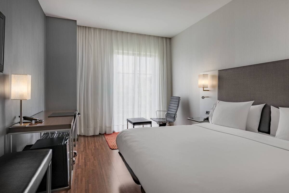 Een moderne hotelkamer met een groot bed, een bureau met lamp, een stoel en een raam met gordijnen.