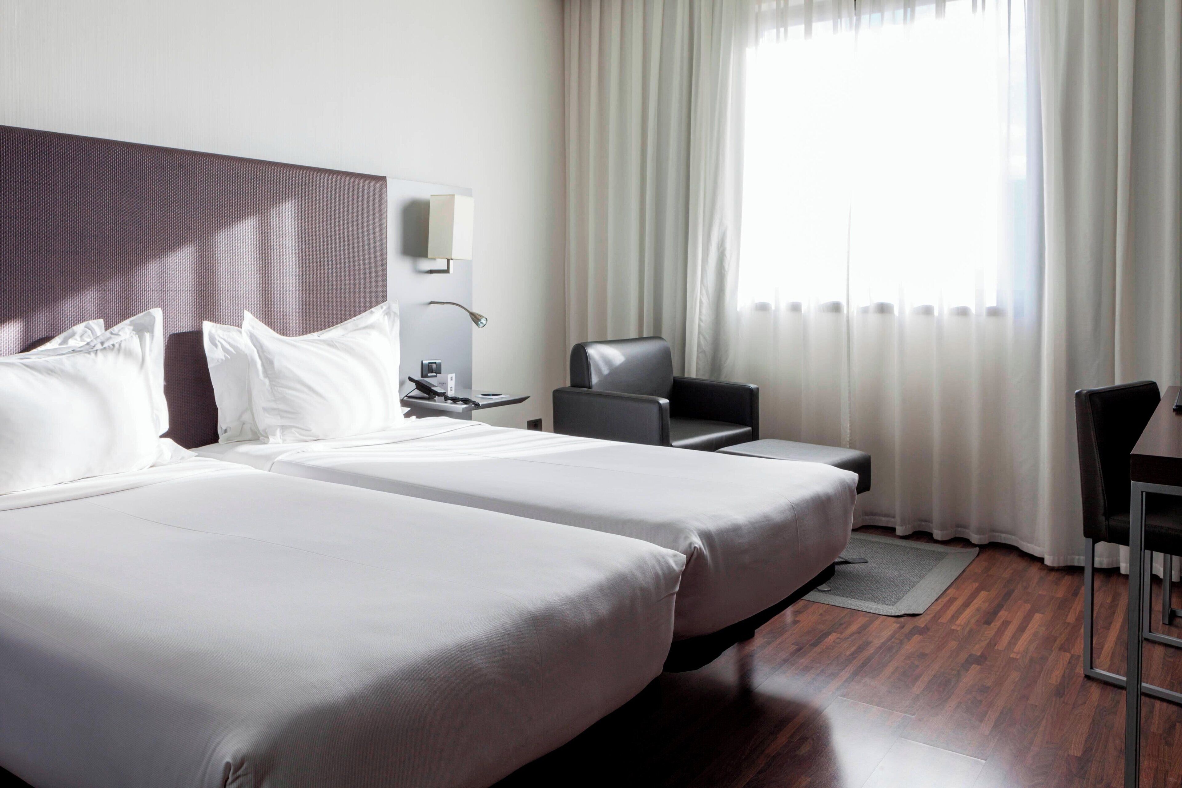 A hotel room with two beds, a desk, a chair, and a large window with curtains.