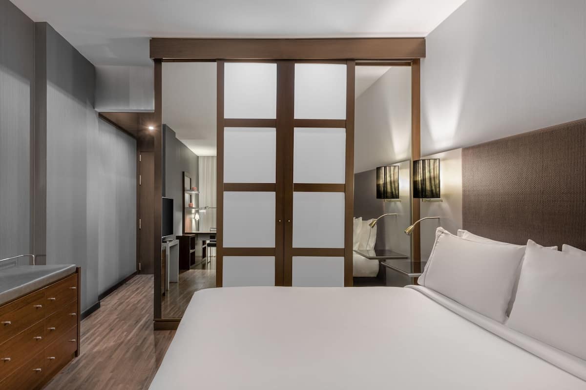 Una habitación de hotel moderna con una cama grande, un armario de madera, un baño con espejo y un escritorio pequeño.