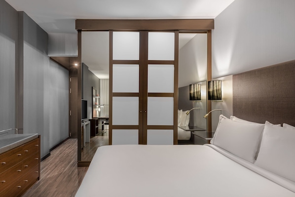A modern hotel room with a large bed, a wooden wardrobe, a bathroom with a mirror, and a small desk.