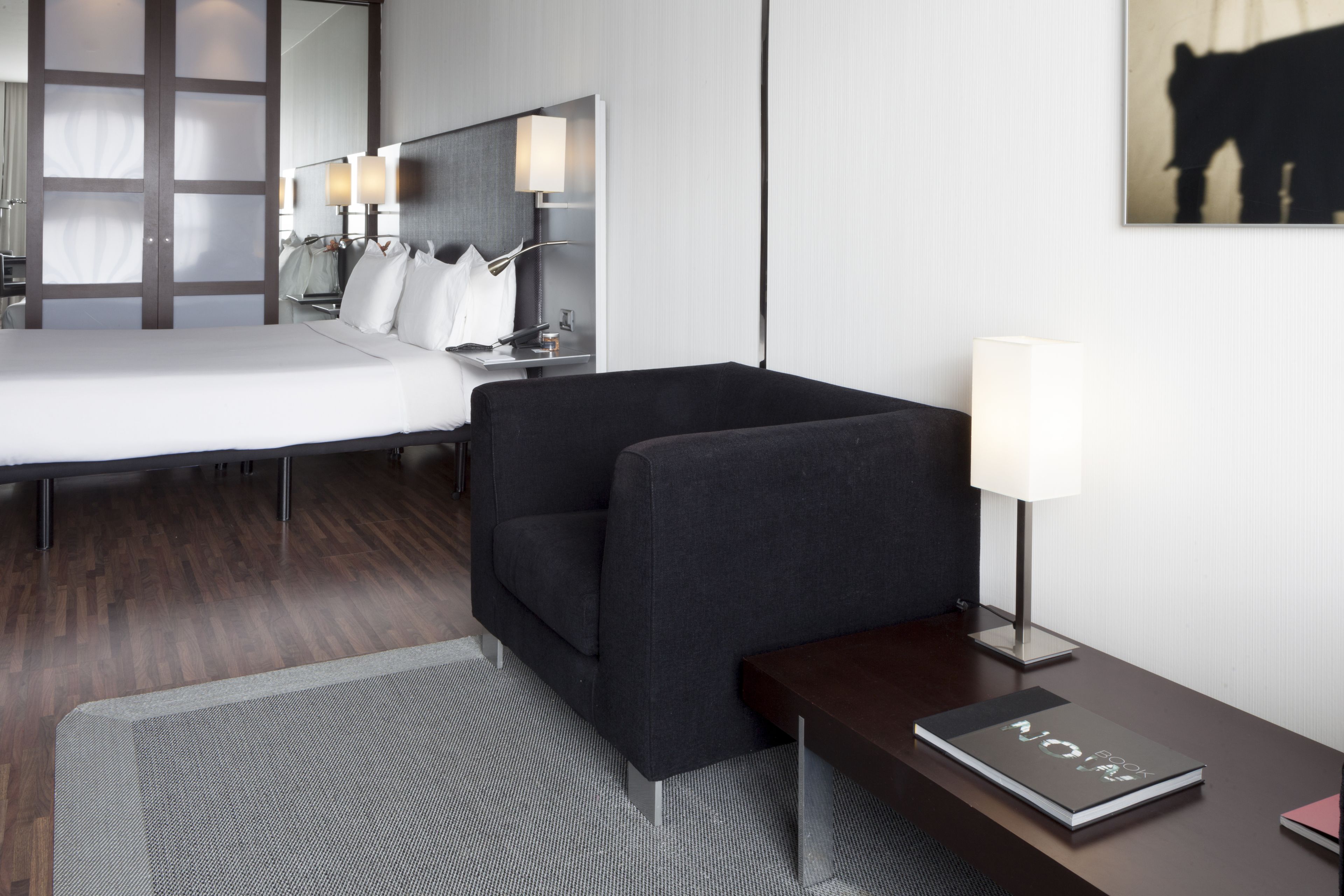 A modern hotel room with a bed, a dark armchair, a bedside table with a lamp, and a book titled "NOVA".