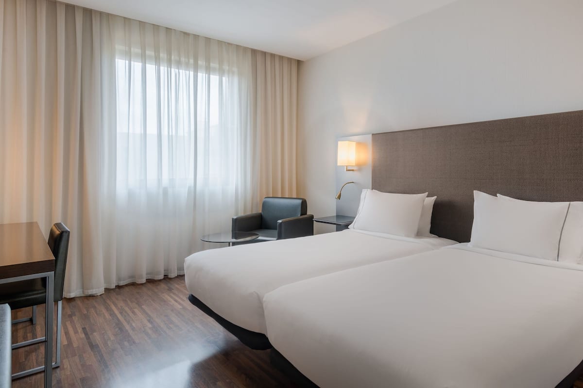 Habitación de hotel con dos camas, un escritorio, una silla, una lámpara y una ventana con cortinas.