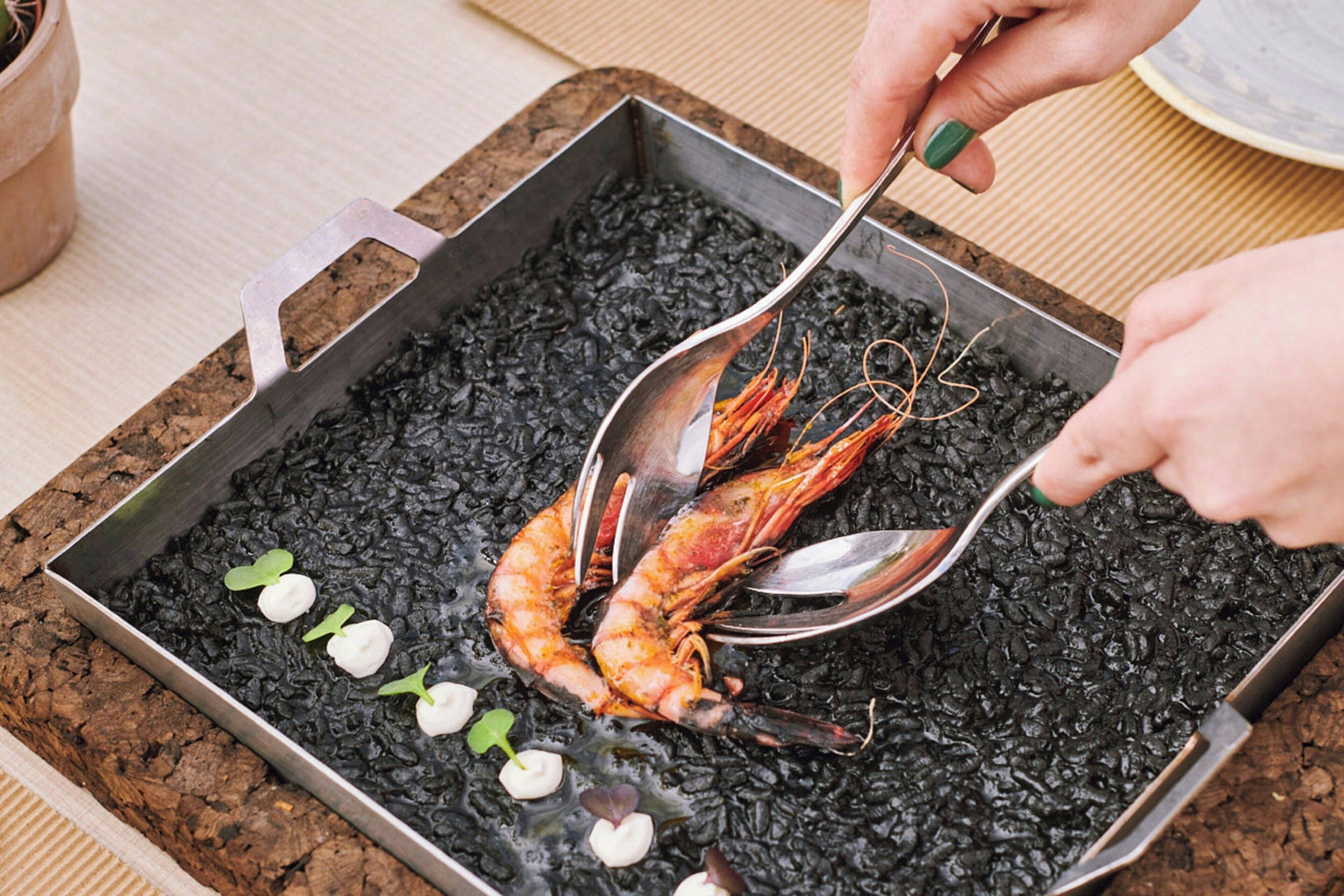 A cooking pan with shrimp on a bed of blackened ingredients, possibly charcoal, with a spoon and small green leaves nearby.