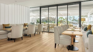 A modern interior with a wooden floor, light-colored furniture, and large windows offering a view of palm trees and a cityscape. - Sabàtic Gavà Mar, Barcelona, a Tribute Portfolio Hotel (Gava)