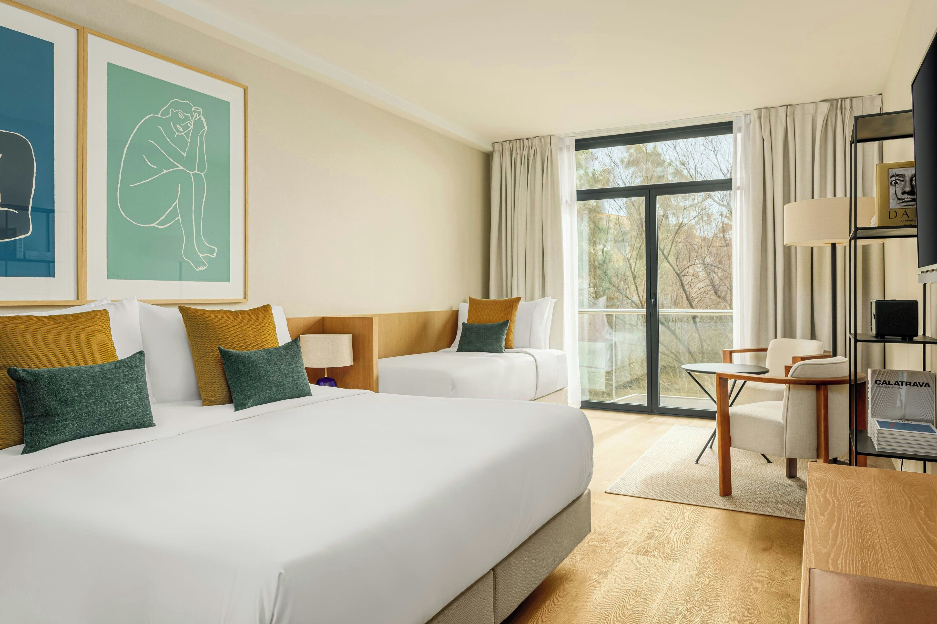 A hotel room with a large bed, two smaller beds, a desk with a chair, a bookshelf, and a large window offering a view of trees.