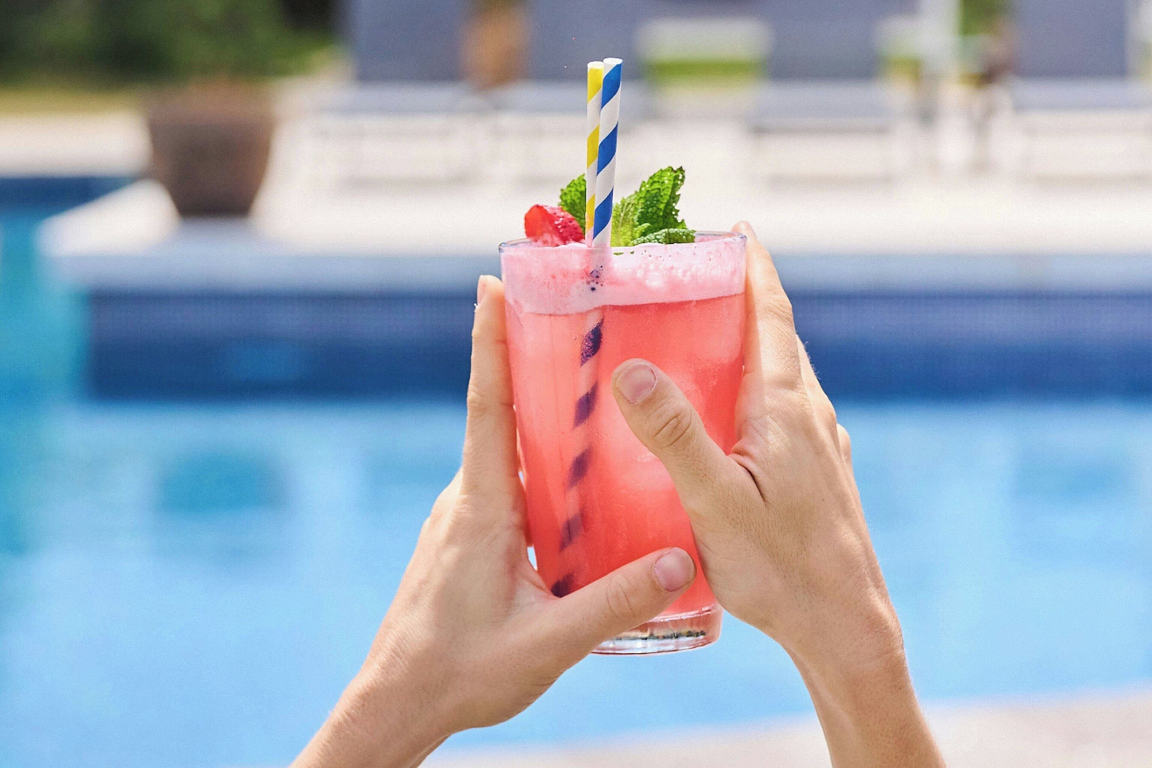 Una persona sosteniendo una bebida rosa con una pajita a rayas, adornada con una hoja de menta y una fresa, junto a una piscina.