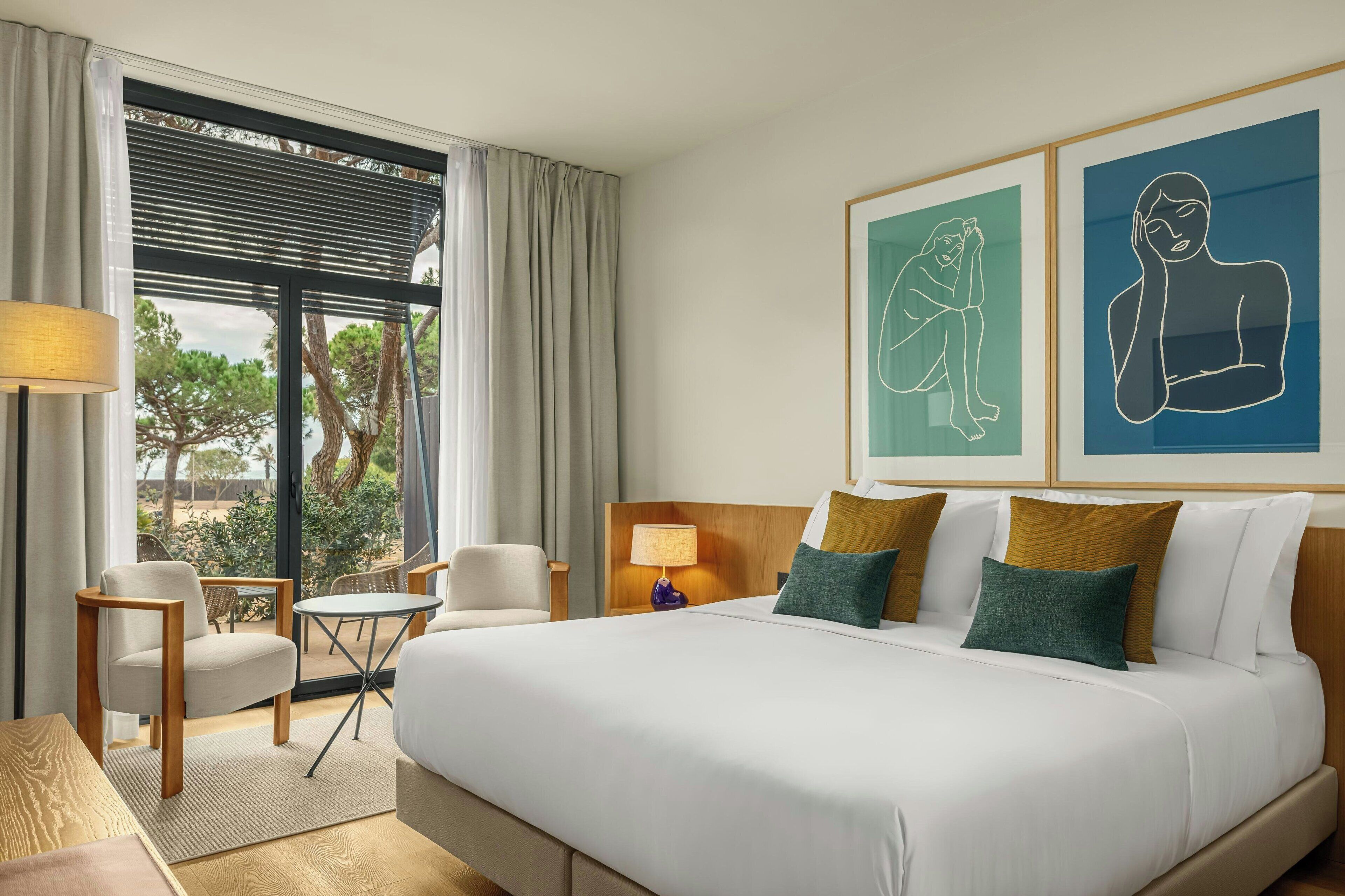 Habitación de hotel con una cama grande, un escritorio, dos sillas, una mesita, una lámpara y obras de arte en la pared.