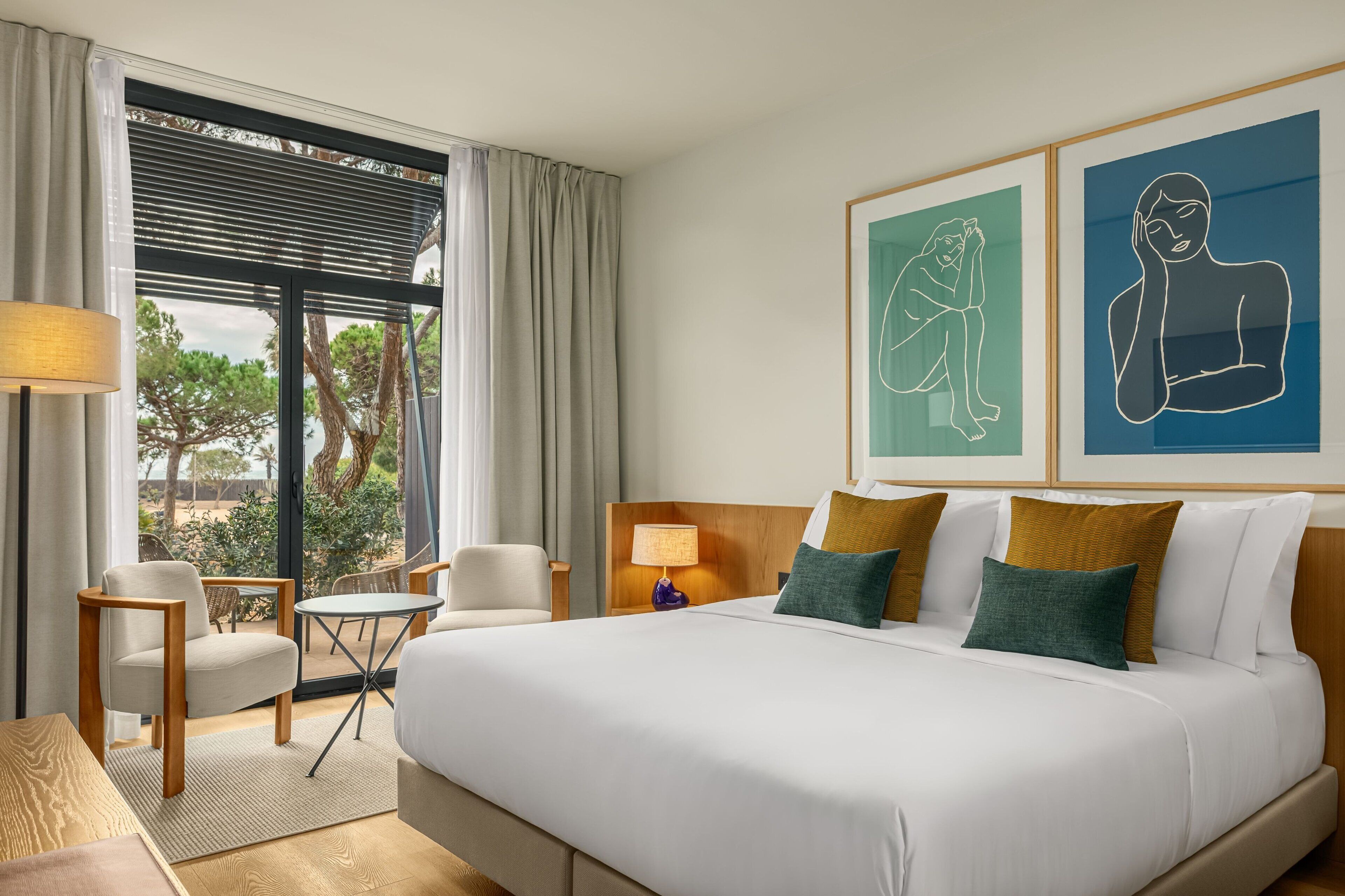 Ein Hotelzimmer mit einem großen Bett, zwei Sesseln, einem kleinen Tisch und einem Fenster mit Vorhängen. An der Wand hängen zwei gerahmte Kunstwerke.