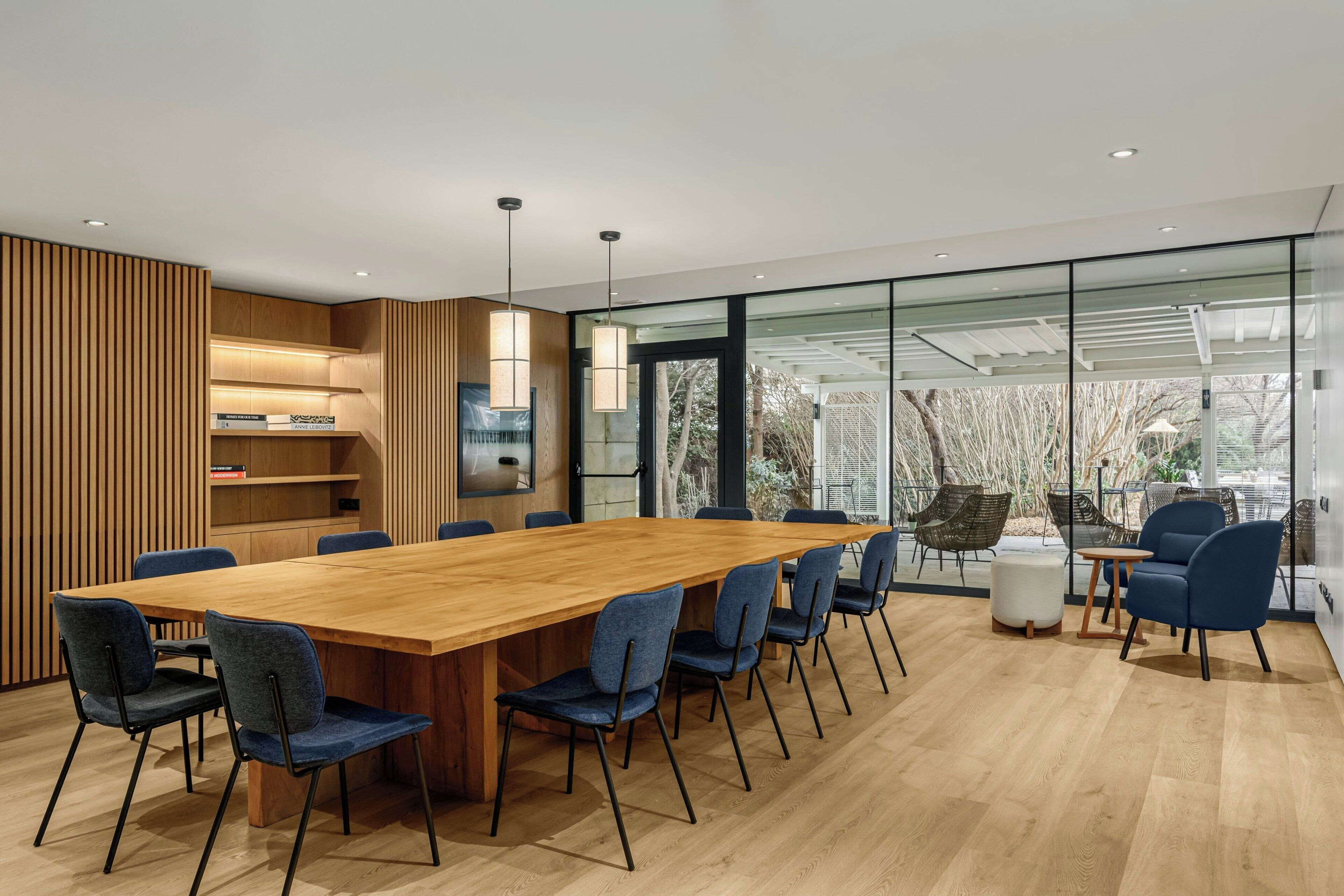 Una sala de reuniones moderna con una larga mesa de madera, sillas tapizadas en azul y un panel de vidrio con vistas a árboles.