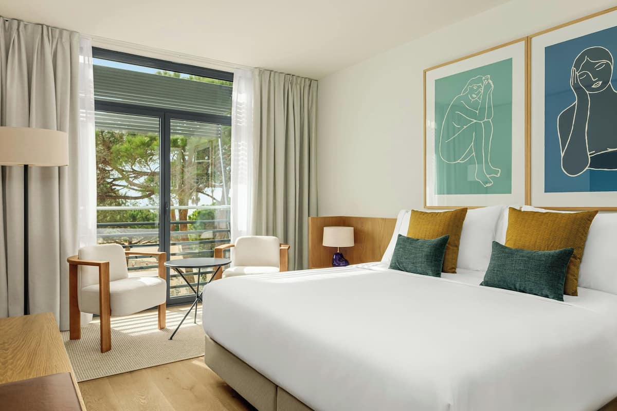 Ein Hotelzimmer mit einem großen Bett, einem Schreibtisch, einem Sessel und Blick ins Freie.