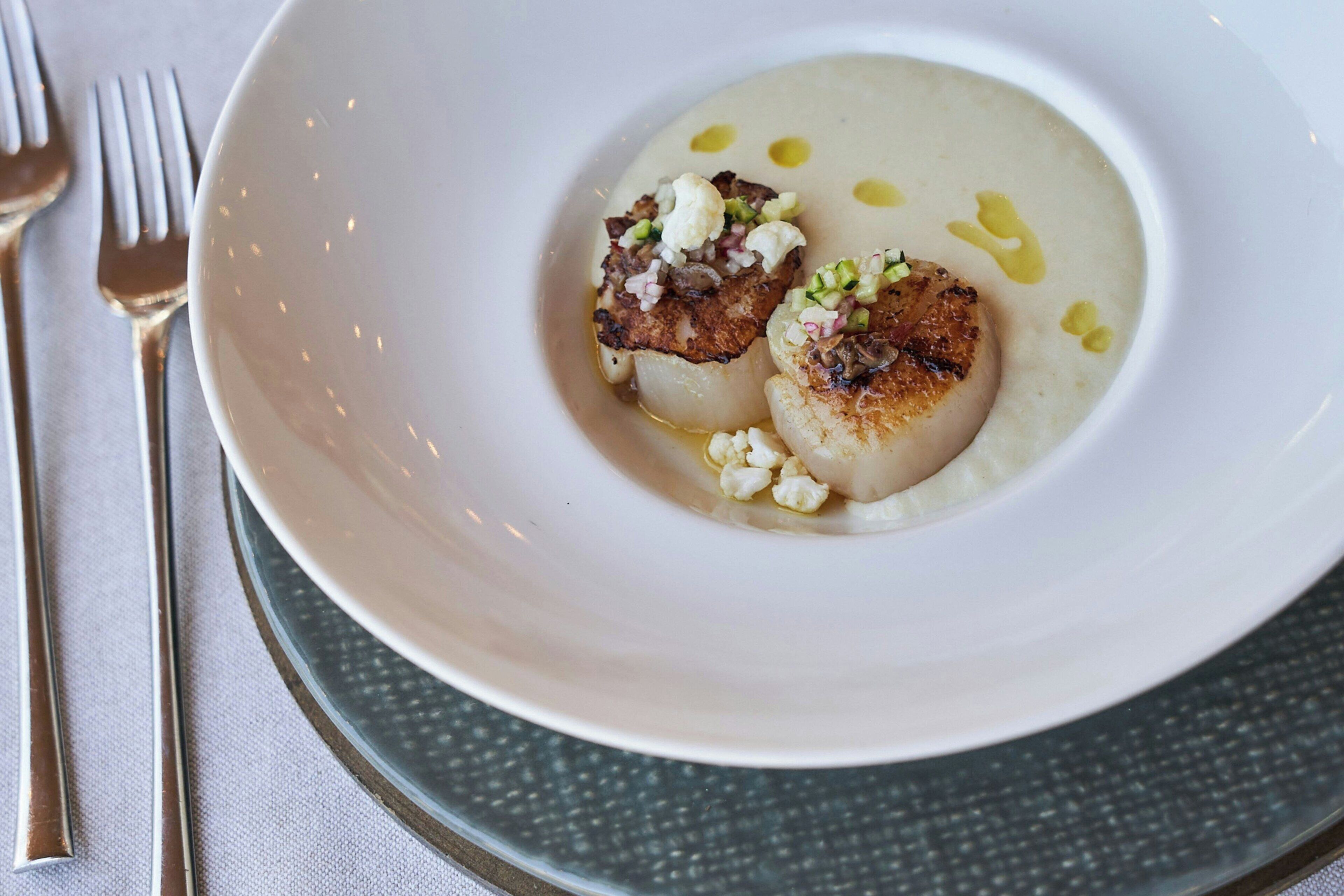 A white plate with a scallop and a creamy puree, accompanied by a fork and knife on a tablecloth.