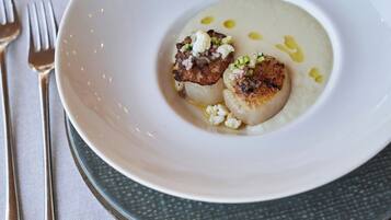 A white plate with a scallop and a creamy puree, accompanied by a fork and knife on a tablecloth.