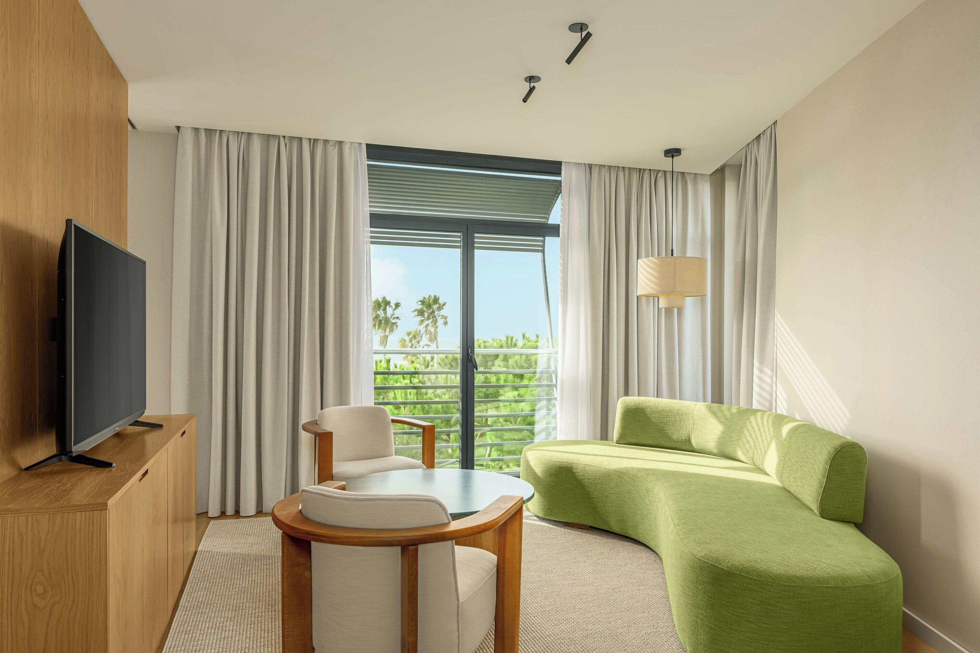 A modern hotel room with a flat-screen TV, a green sofa, a white armchair, and a small table.