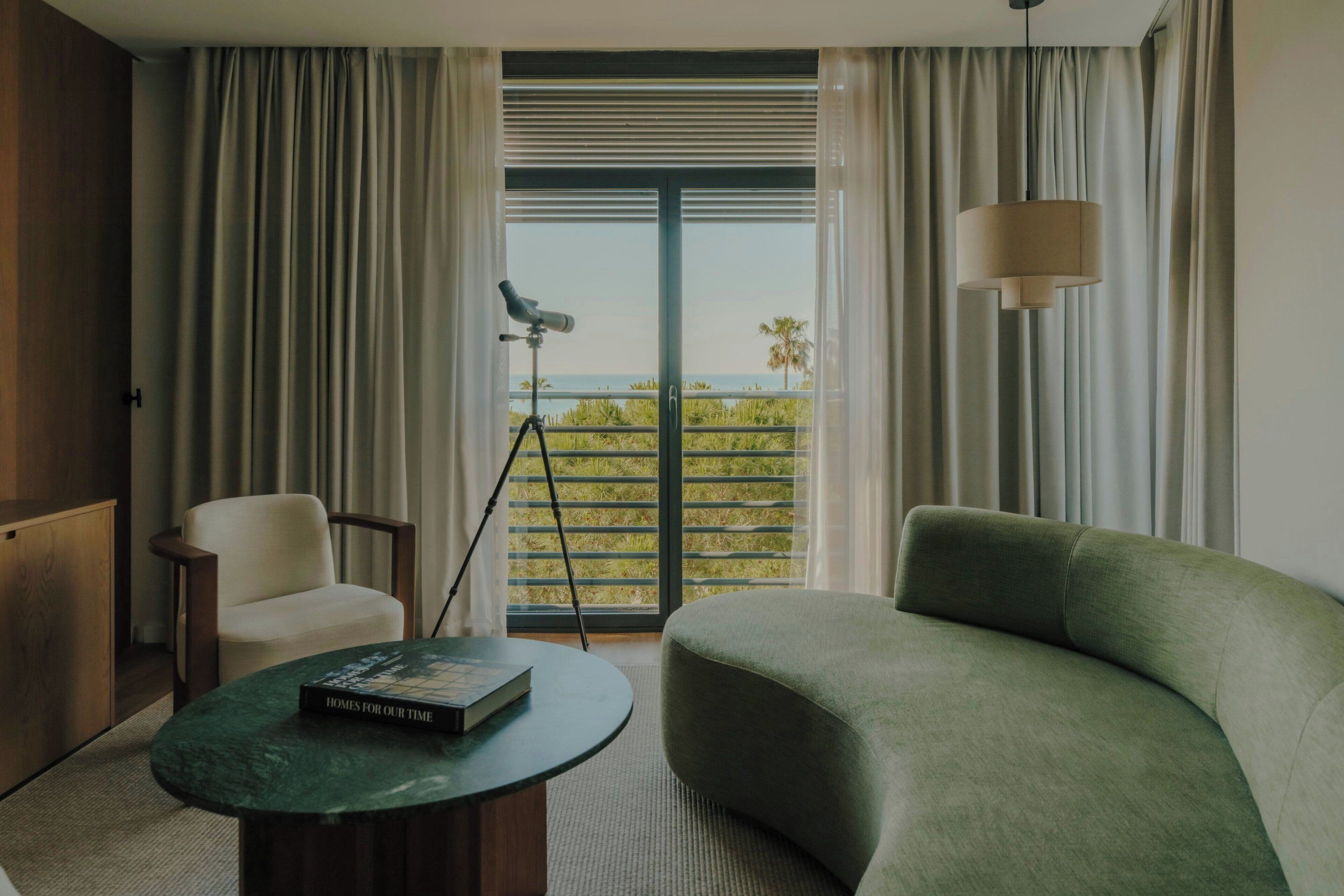 A modern hotel room with a green sofa, a small round table with a book, a tripod with a camera, and a view of the sea through large windows.
