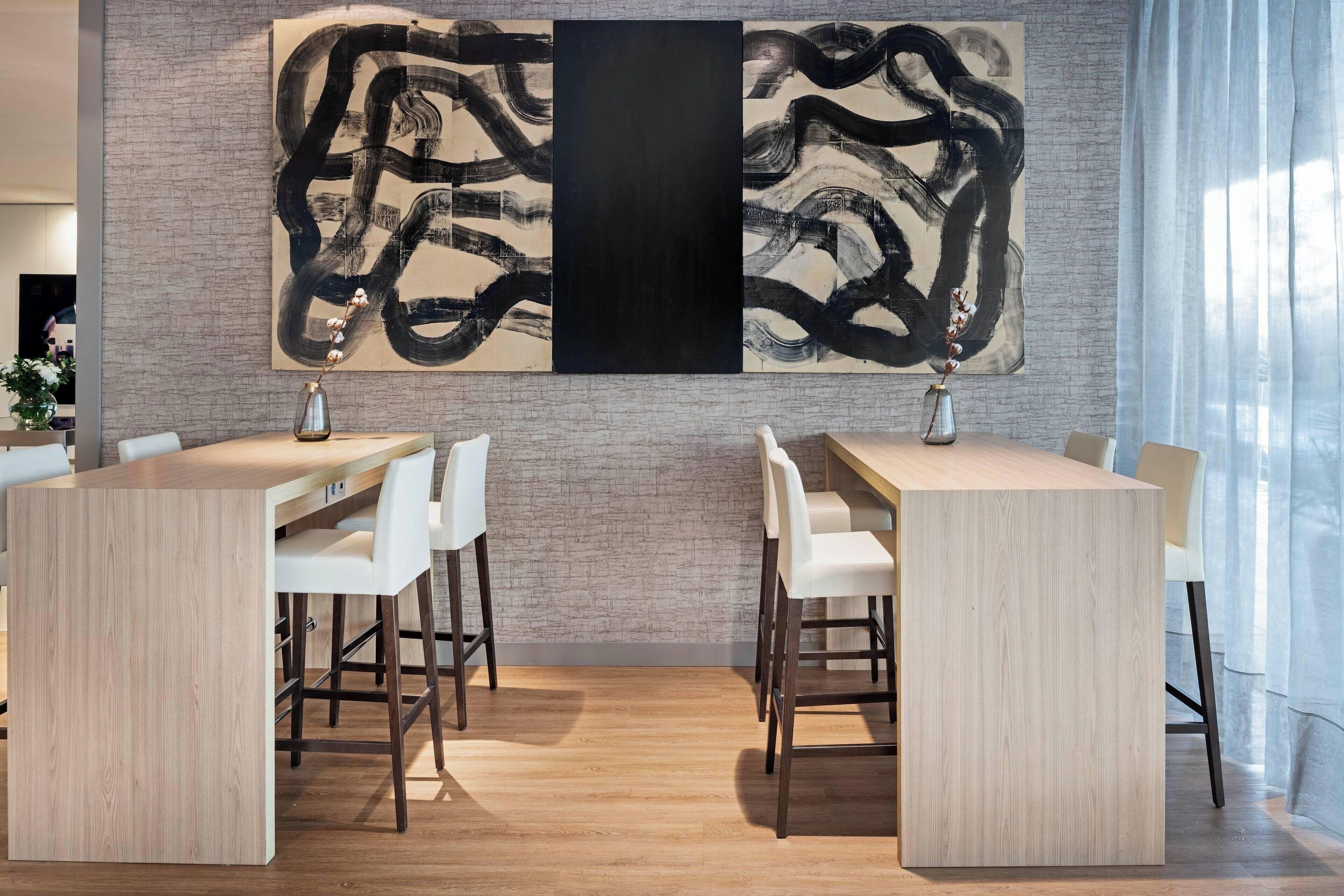 Un espace repas moderne avec deux tables en bois et des chaises blanches, une grande peinture abstraite au mur et un mur en briques.