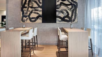 Un área de comedor moderna con dos mesas de madera y sillas blancas, un cuadro abstracto grande en la pared y un muro de ladrillo.