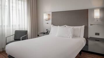 Una habitación de hotel con una cama grande, una silla, una mesita de noche y una lámpara fijada a la pared.
