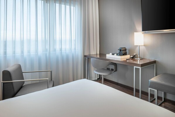 A hotel room with a bed, a desk with a lamp, a chair, and a TV mounted on the wall.