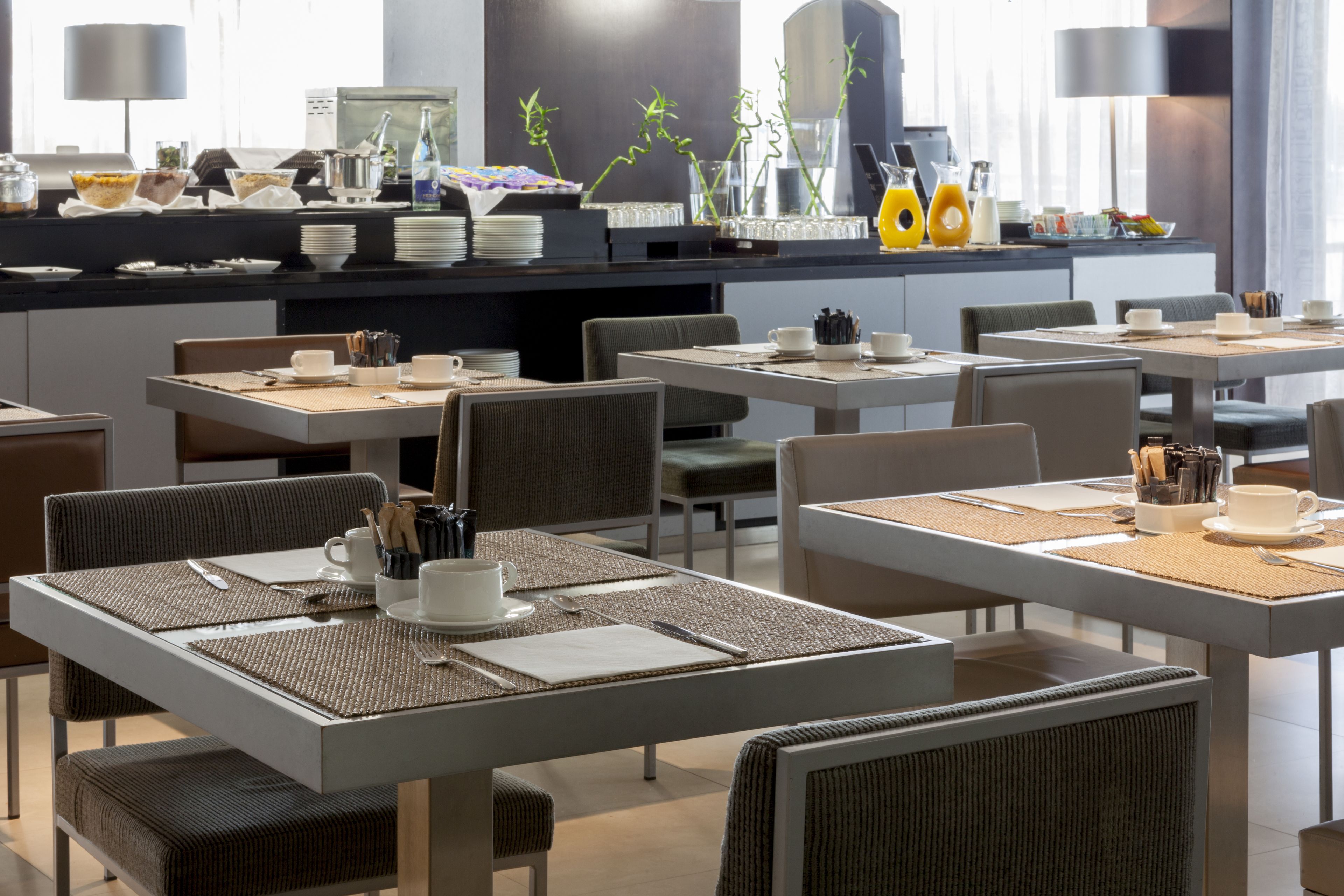 Un comedor de hotel moderno con varias mesas y sillas, un mostrador de buffet con distintos alimentos, y un diseño contemporáneo y limpio.