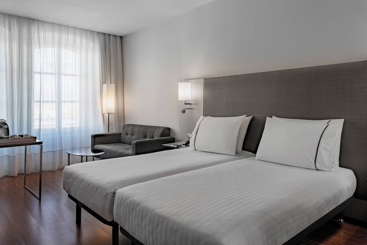 Une chambre d’hôtel avec deux lits, un bureau, une chaise, une lampe et une vue sur l’extérieur.