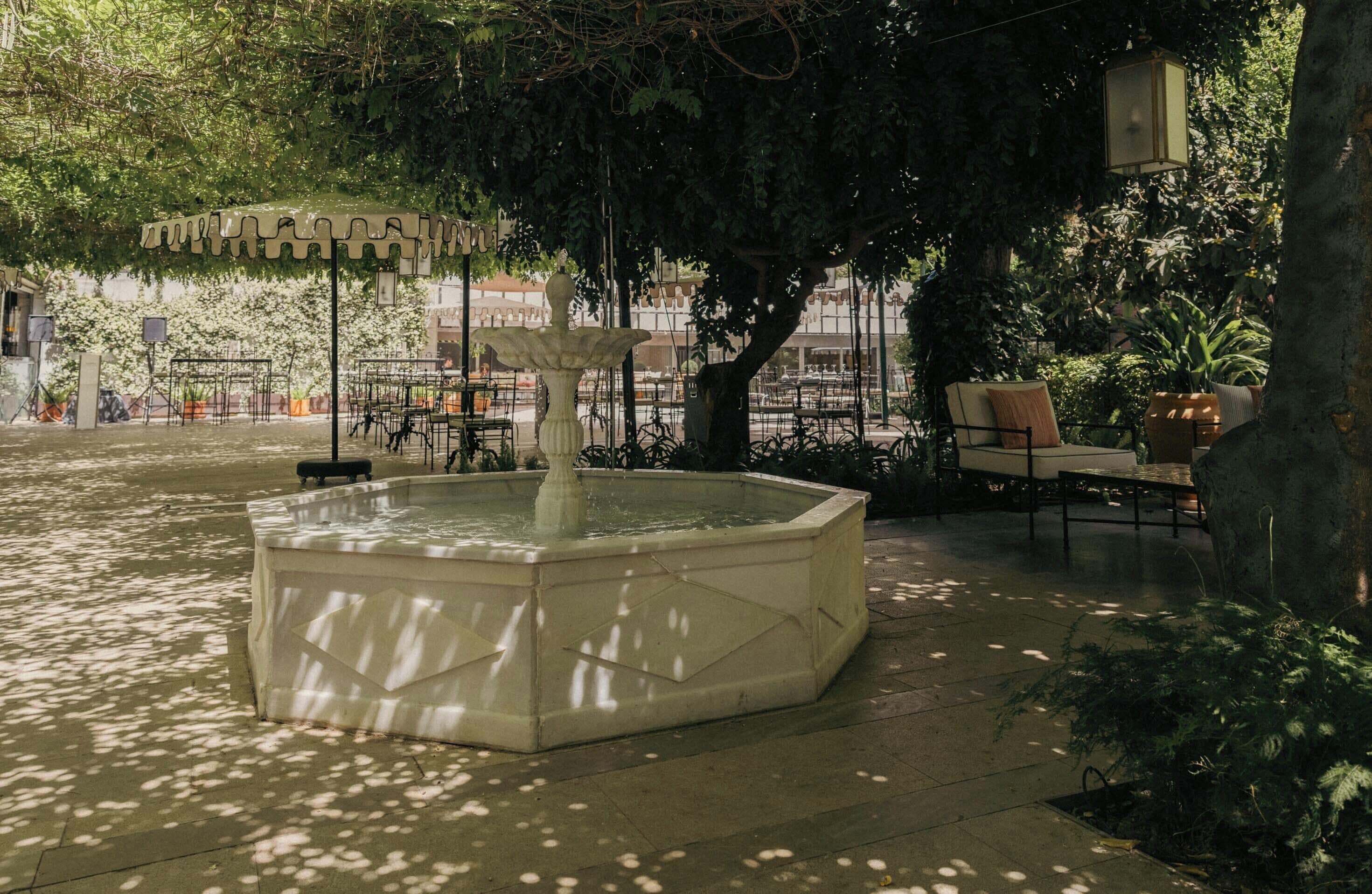 Une fontaine extérieure de forme carrée, dotée d’une ouverture centrale, et entourée d’une zone pavée avec des chaises et des tables.