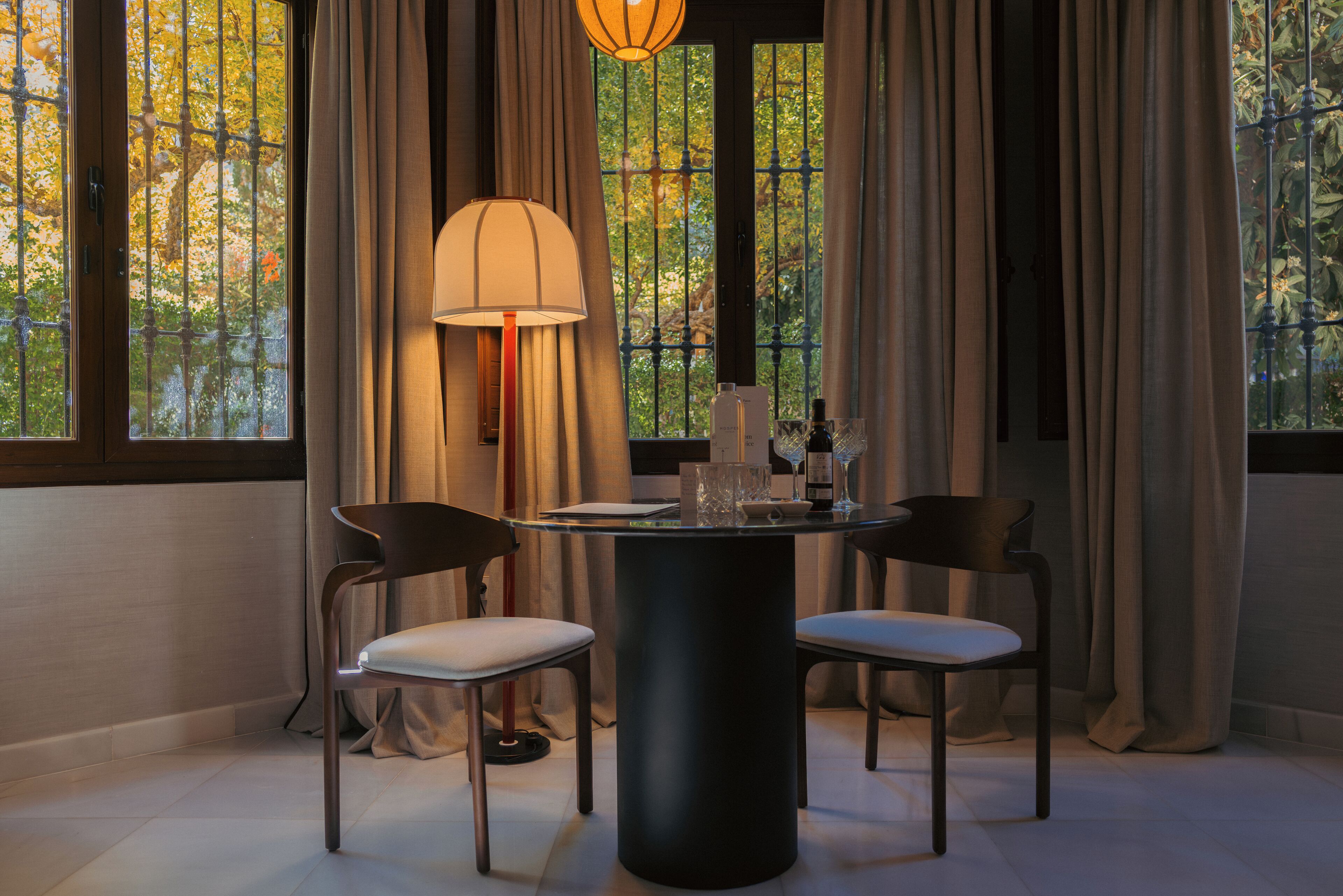 A dining area with a round table, two chairs, a lamp, and a view of trees through large windows.