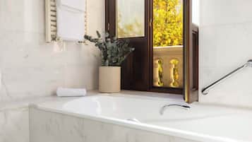 Un baño con una bañera de mármol blanco, una planta en maceta, una toalla y vistas a un balcón con follaje amarillo.