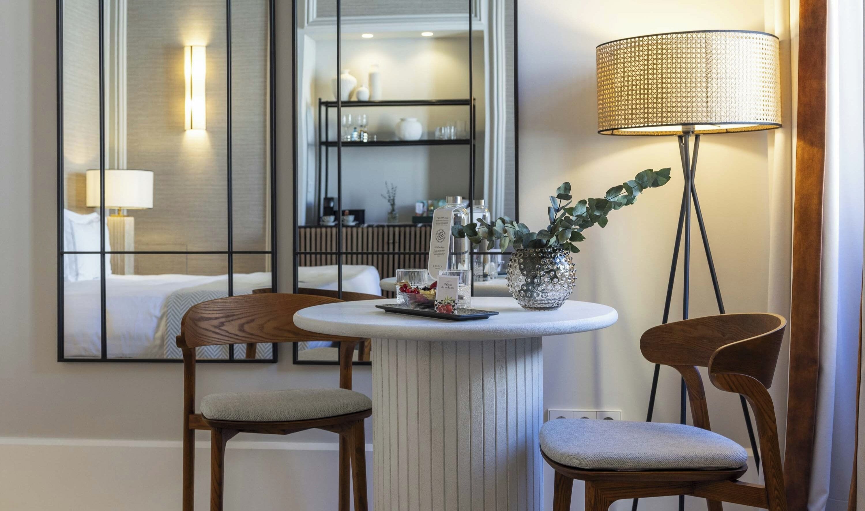 A hotel room with a round table, two wooden chairs, a floor lamp, a mirror, and a shelf with toiletries.