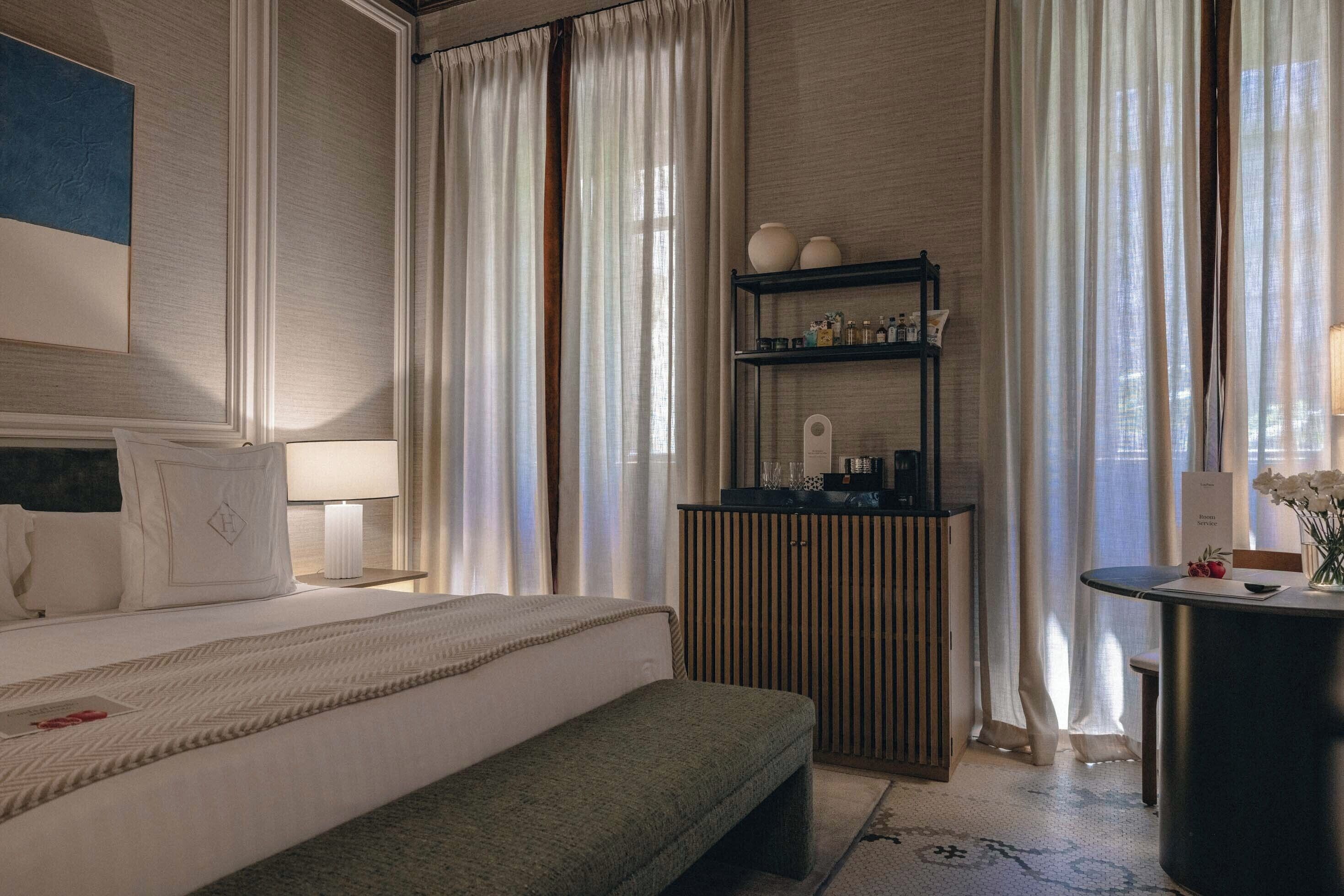 A hotel room with a bed, bedside table, lamp, curtains, a shelf with toiletries, and a small table with flowers.