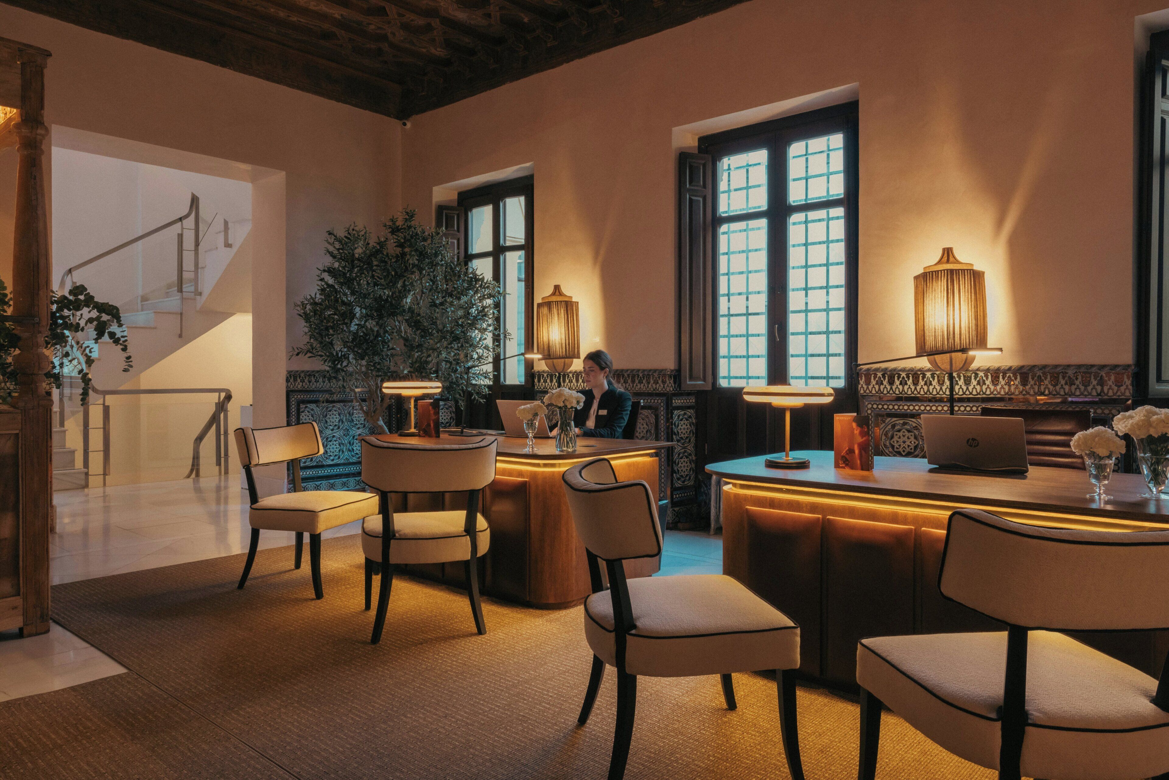 A hotel lobby with a staircase, a reception desk, multiple seating areas, and large windows.