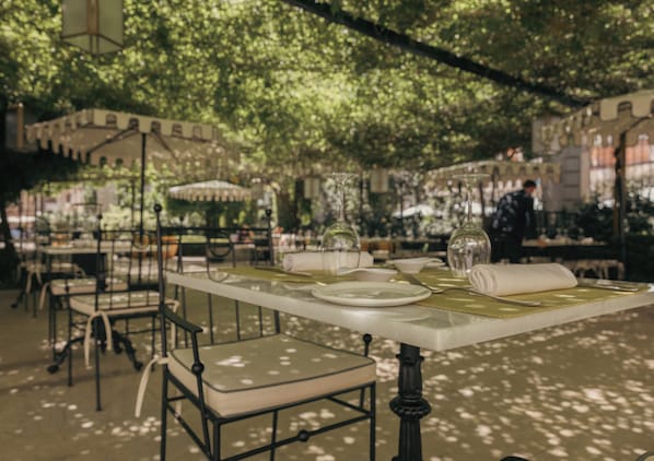 An outdoor dining area with tables set for two, surrounded by greenery and architectural elements.