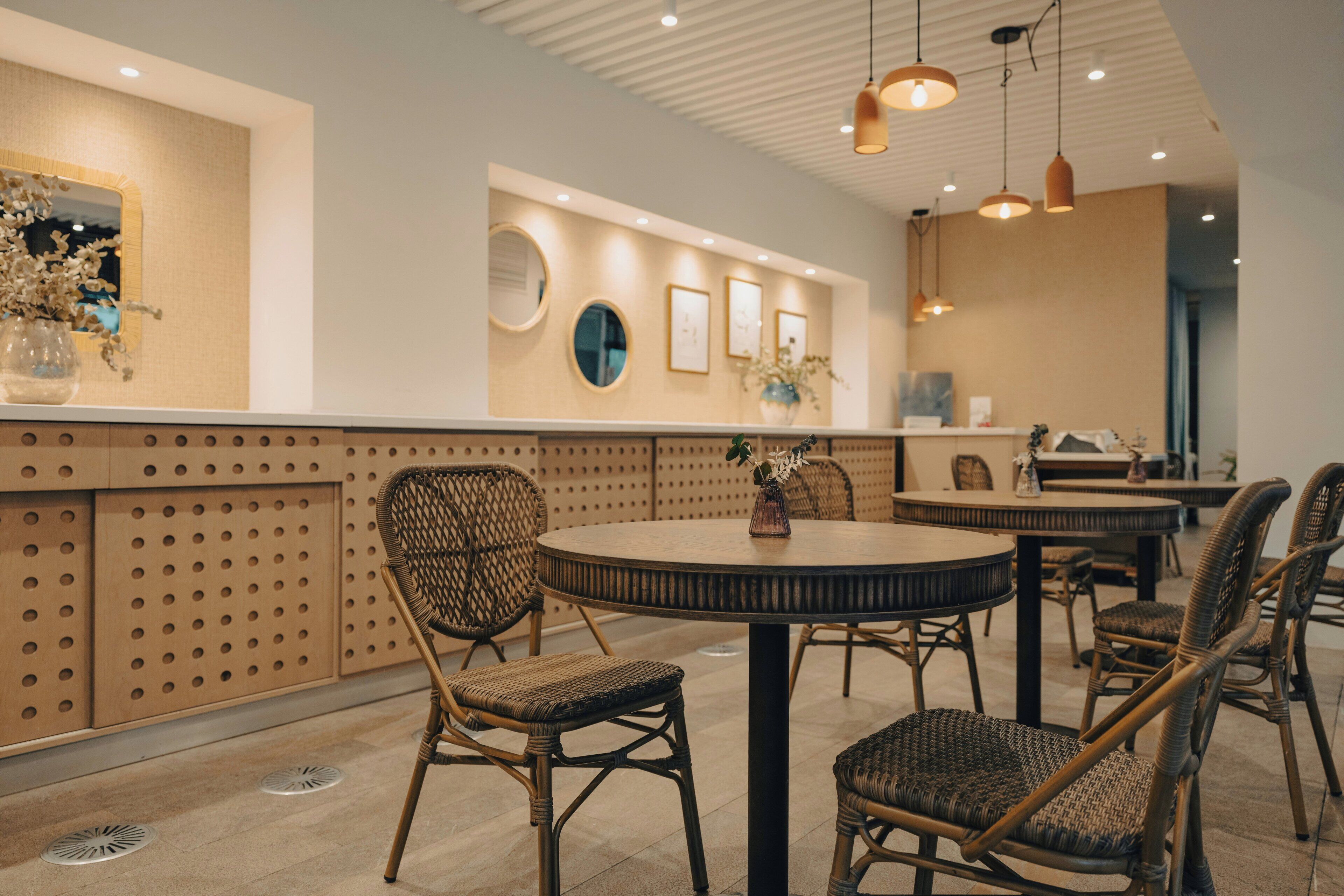 L’intérieur d’un restaurant avec des tables rondes, des chaises en osier et un mur orné de miroirs circulaires et d’œuvres d’art encadrées.