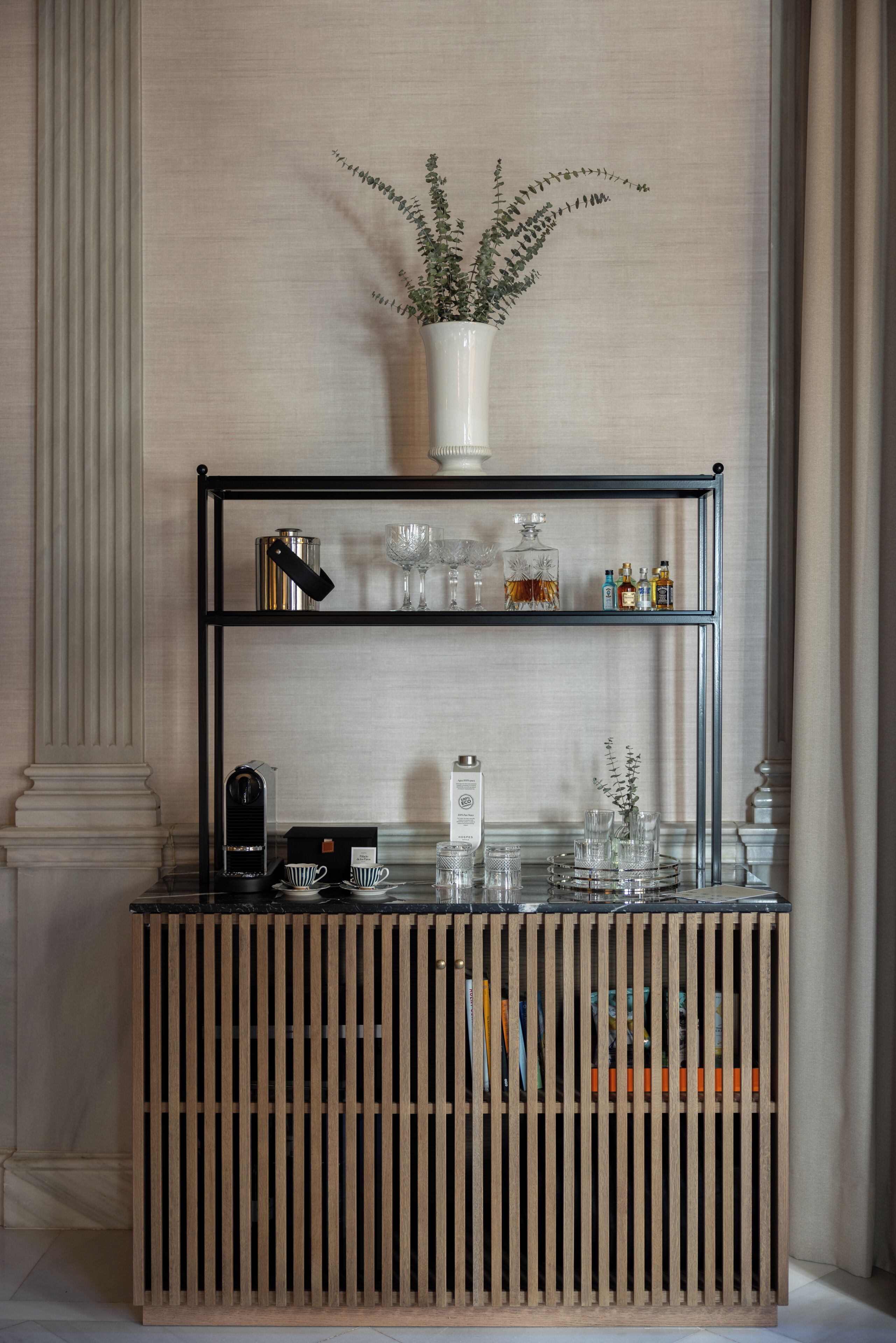 A bar area with a wooden slatted base, a metal shelf with glassware, a potted plant, and a coffee machine.
