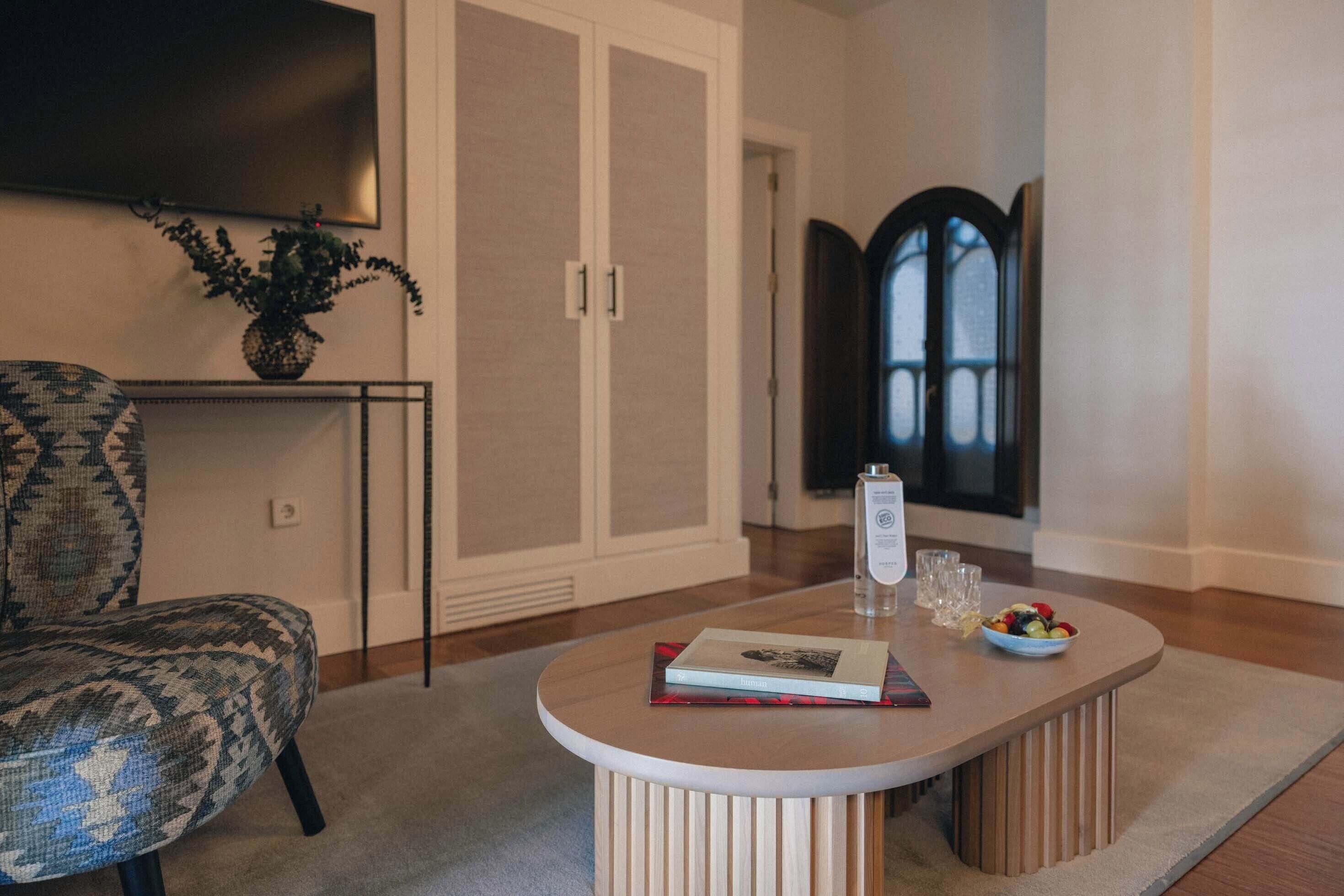 Un salón con un sillón estampado, una mesa redonda con un libro y un cuenco de fruta, un televisor de pantalla plana y una planta en maceta.