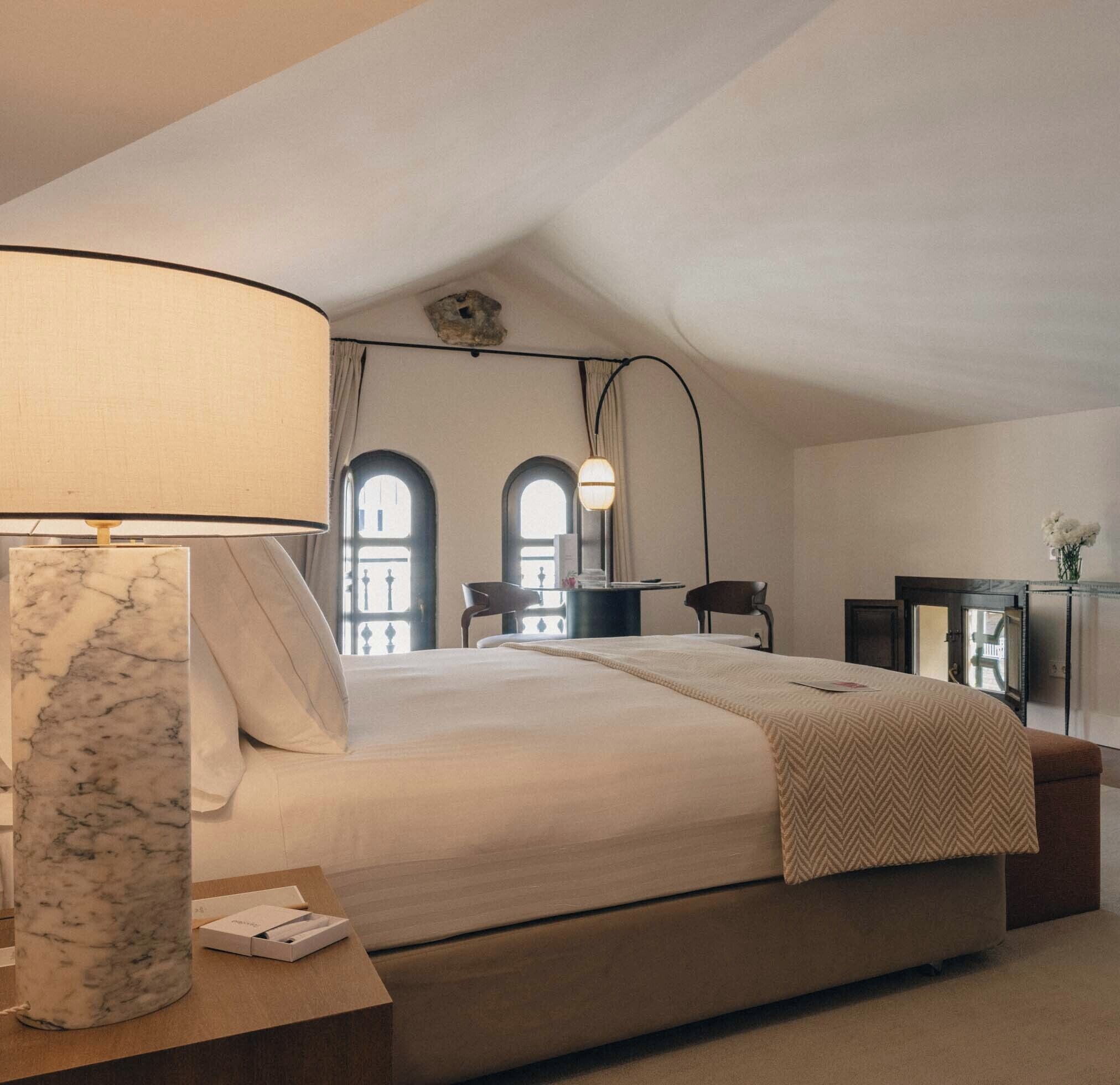 A bedroom with a large bed, a marble bedside table, a desk, two chairs, and a lamp.