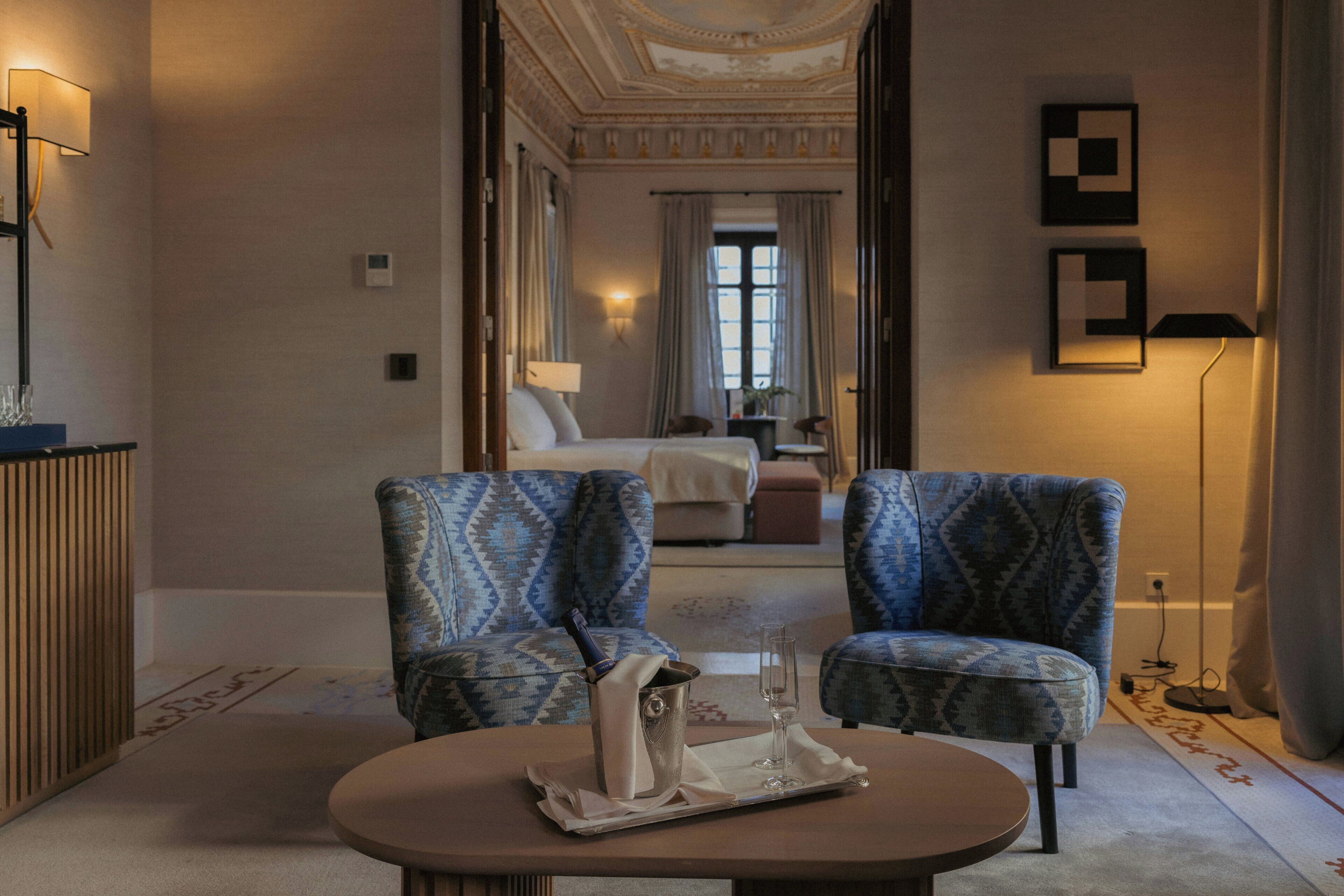 A hotel room with a bed, two patterned armchairs, a small table with a bottle and glasses, and a lamp.