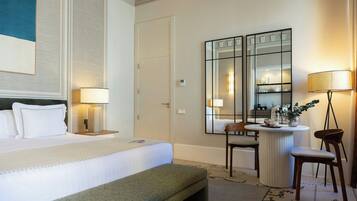 A hotel room with a bed, bedside lamps, a bench, a small table with a chair, a mirror, and a floor lamp.