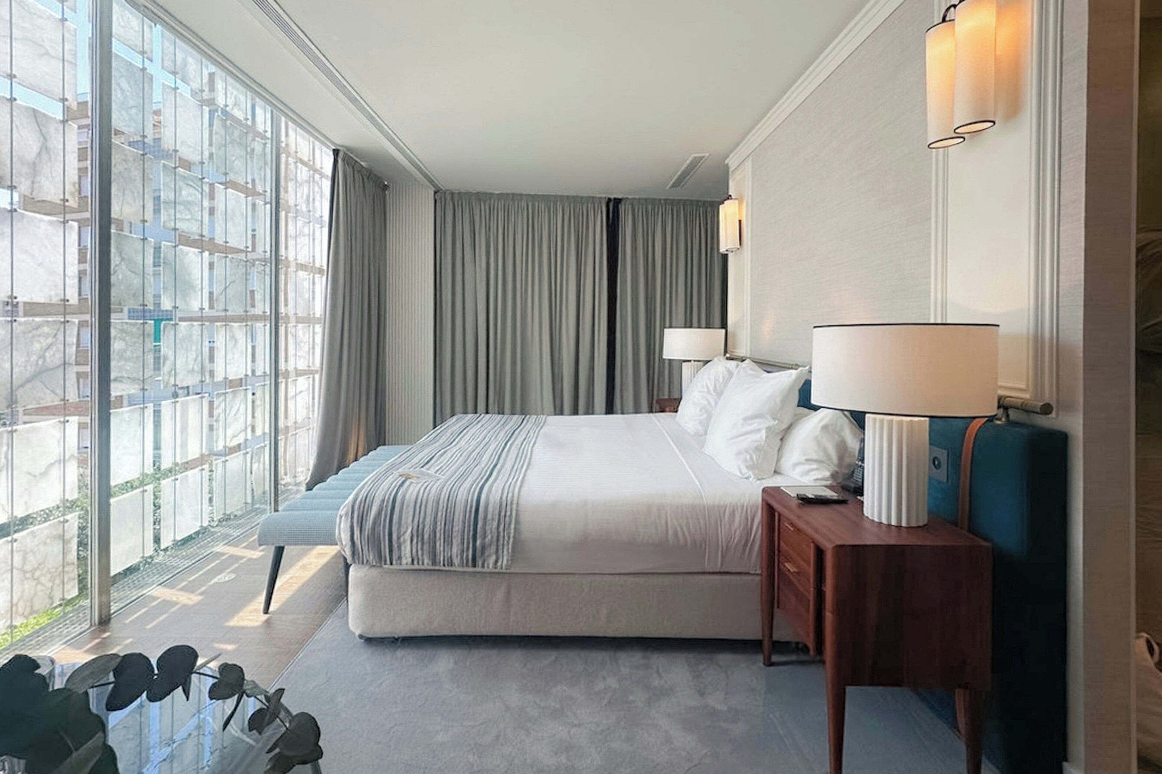 A hotel room with a large bed, a bedside table with a lamp, a window with curtains, and a view of the outside.